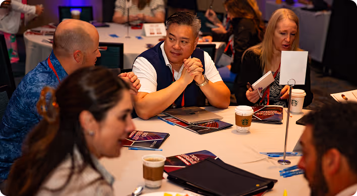 Group of professionals engaged in discussion around a round table with notebooks, pens, and coffee cups during a conference or meeting.