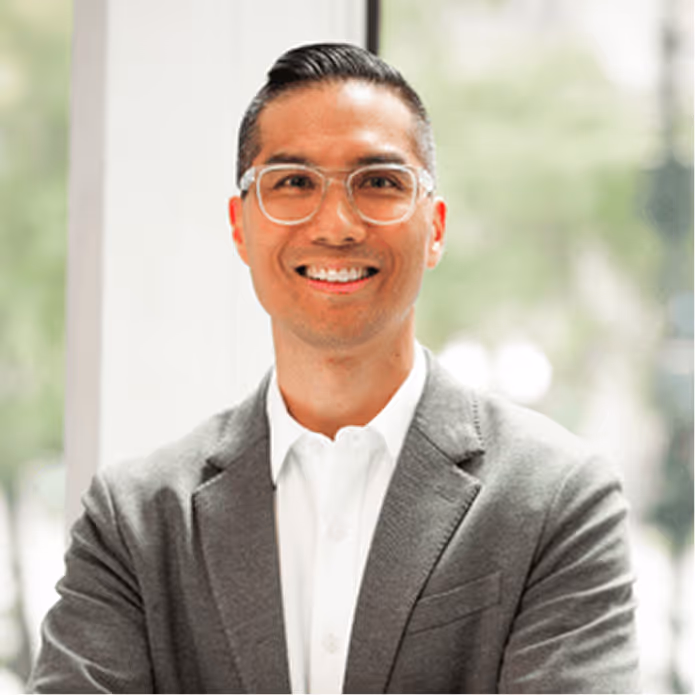 Smiling man wearing glasses, a white shirt, and a gray blazer standing in front of a bright window.