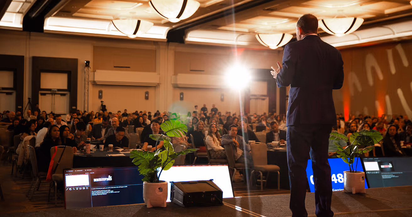 Man in a suit speaking on stage to a large audience seated at round tables in a conference room.