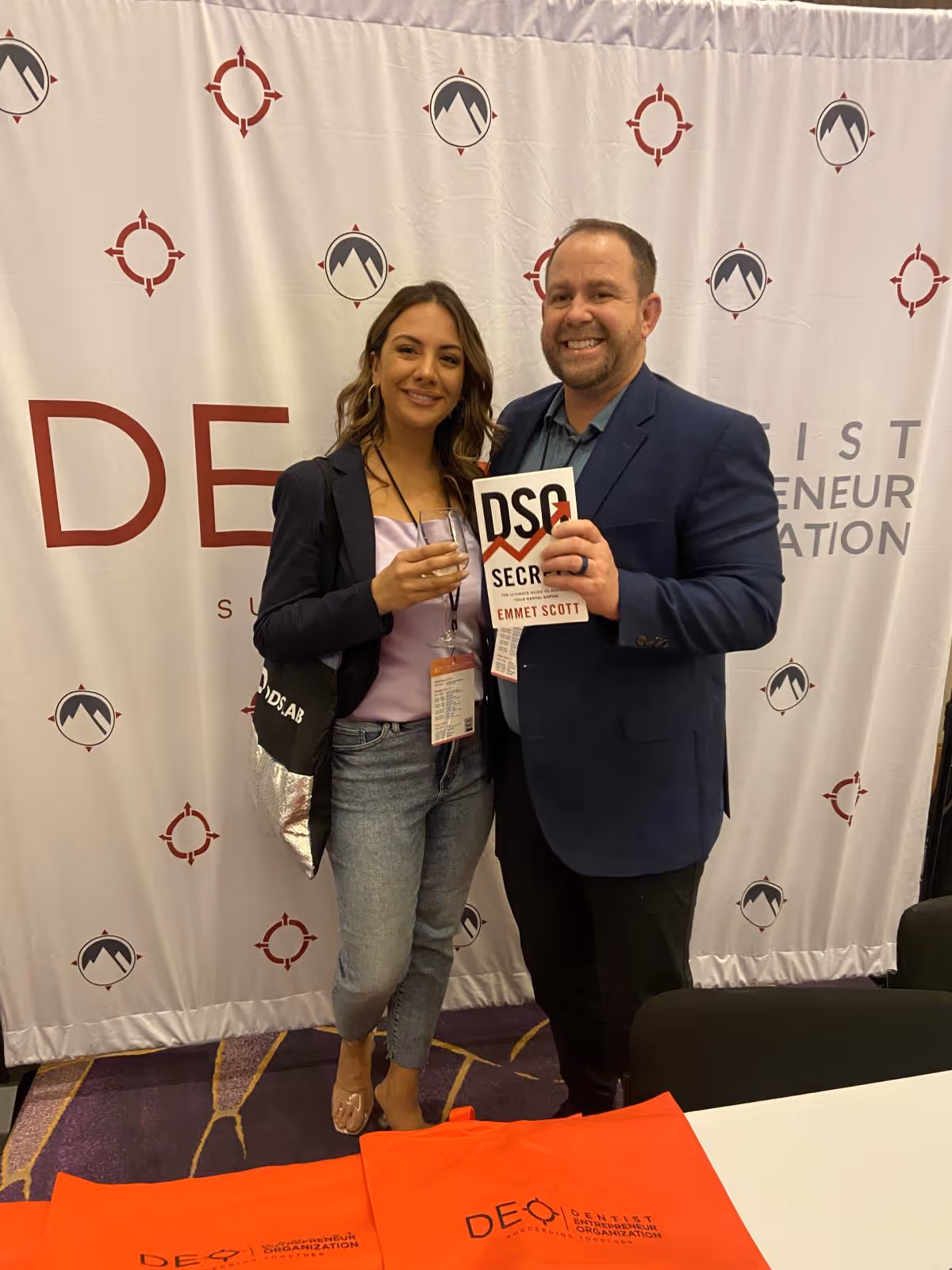 Smiling man and woman standing together at a Dentist Entrepreneur Organization event, the man holding the book 'DSO Secrets' by Emmet Scott.