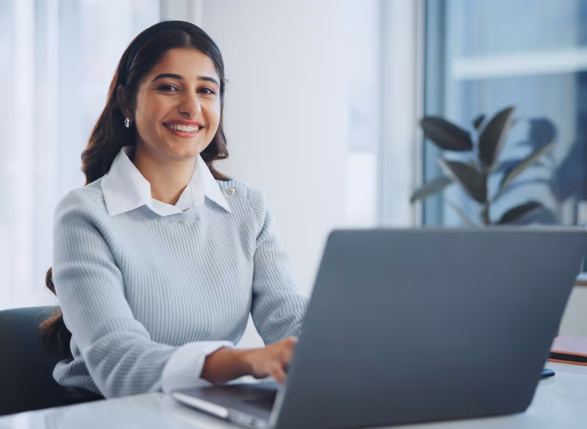 Smiling woman with dark hair wearing a light gray sweater working on a laptop in a bright office.