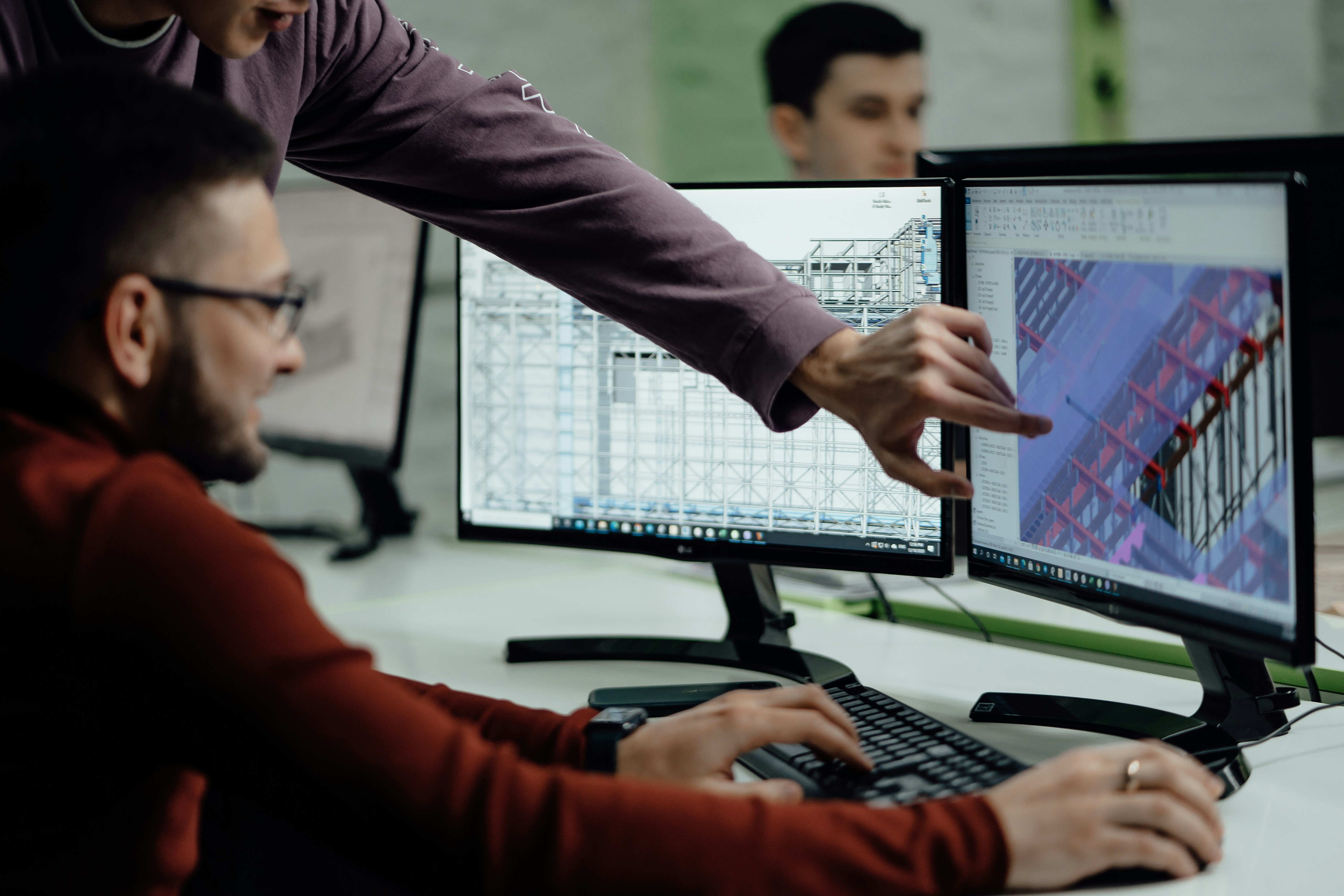 Two men working on 3D architectural designs displayed on dual computer monitors in an office.