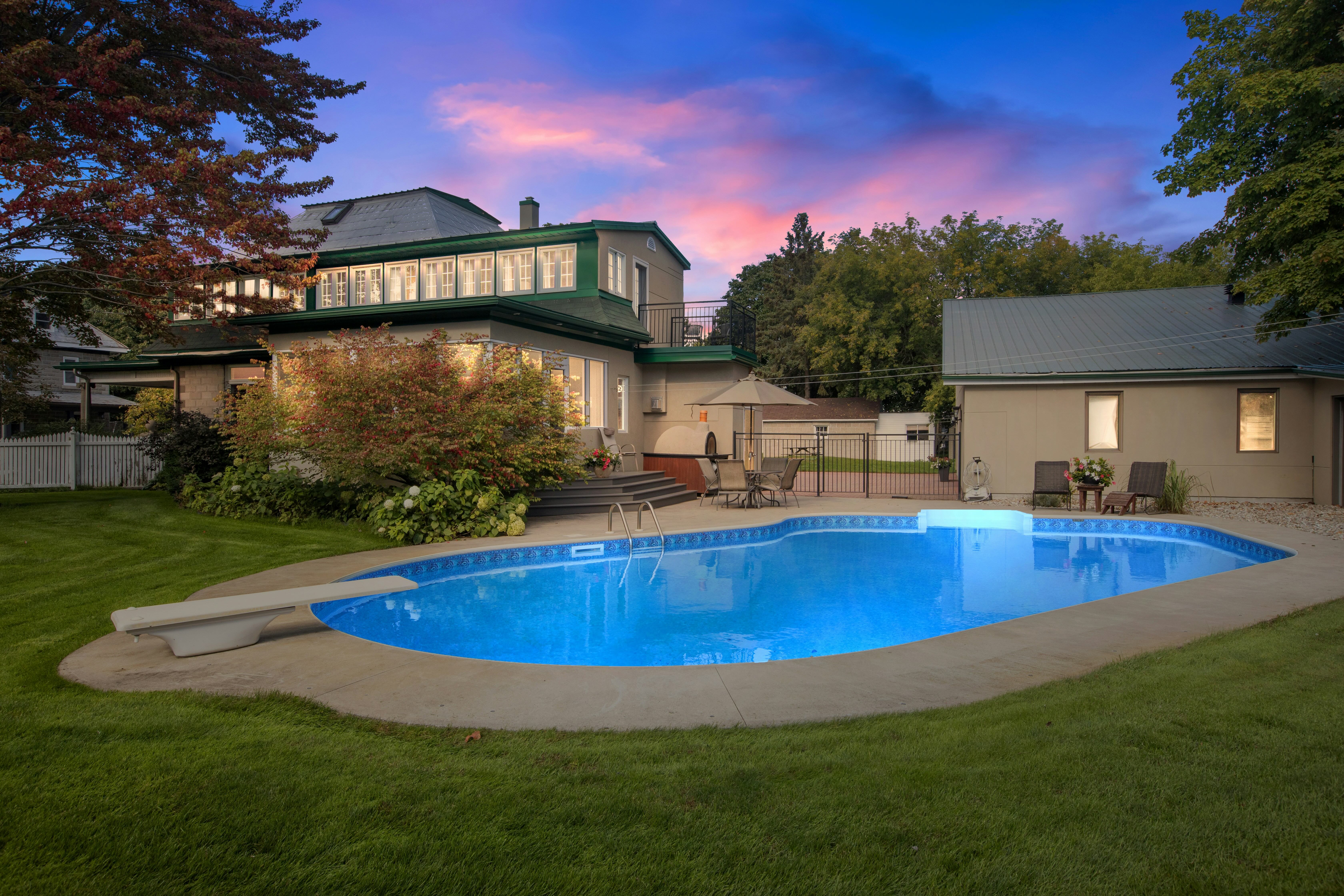 Backyard with illuminated swimming pool, diving board, lawn, patio furniture, and a two-story house at sunset.