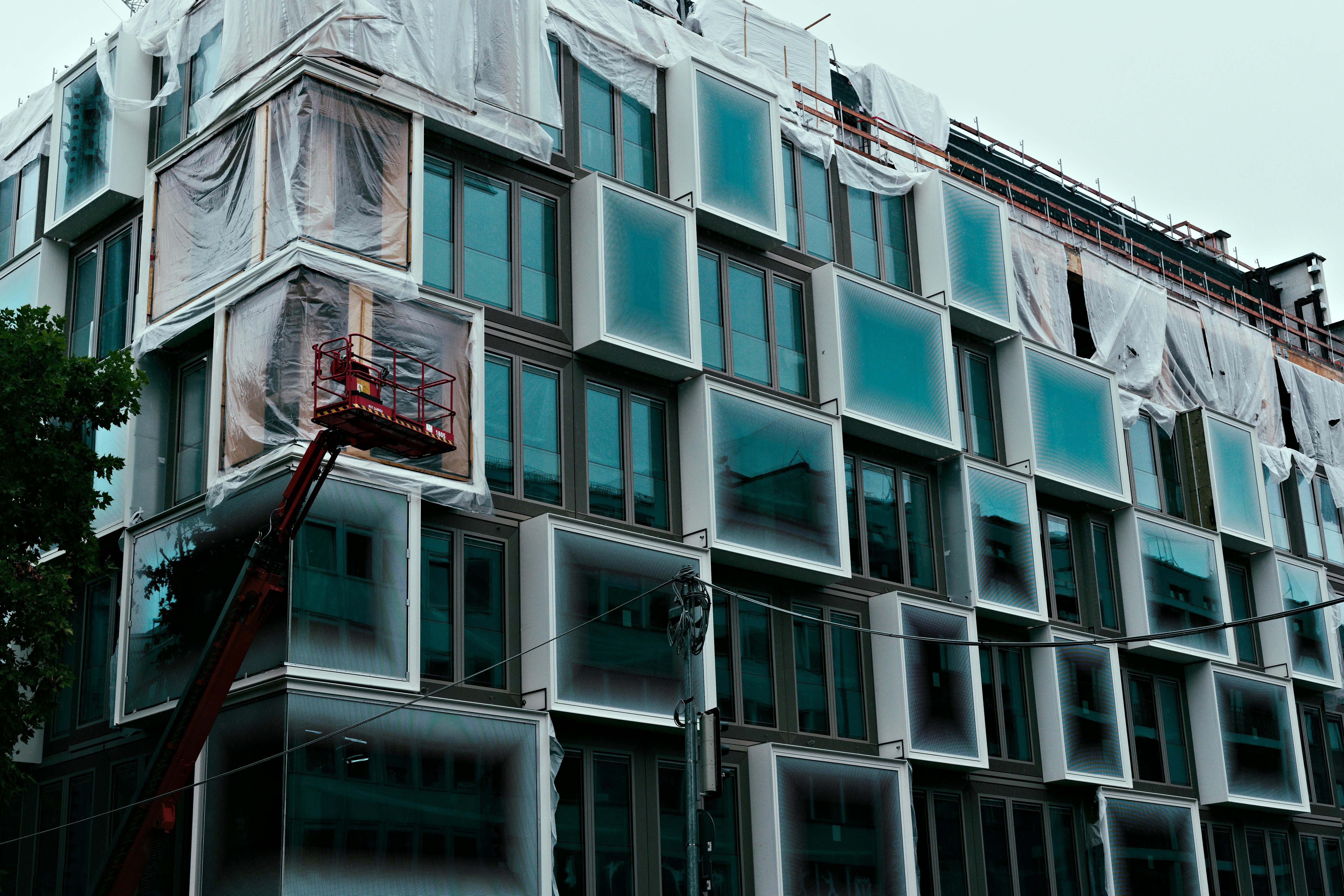 Building facade under construction with large blue glass windows and a lift platform in front.