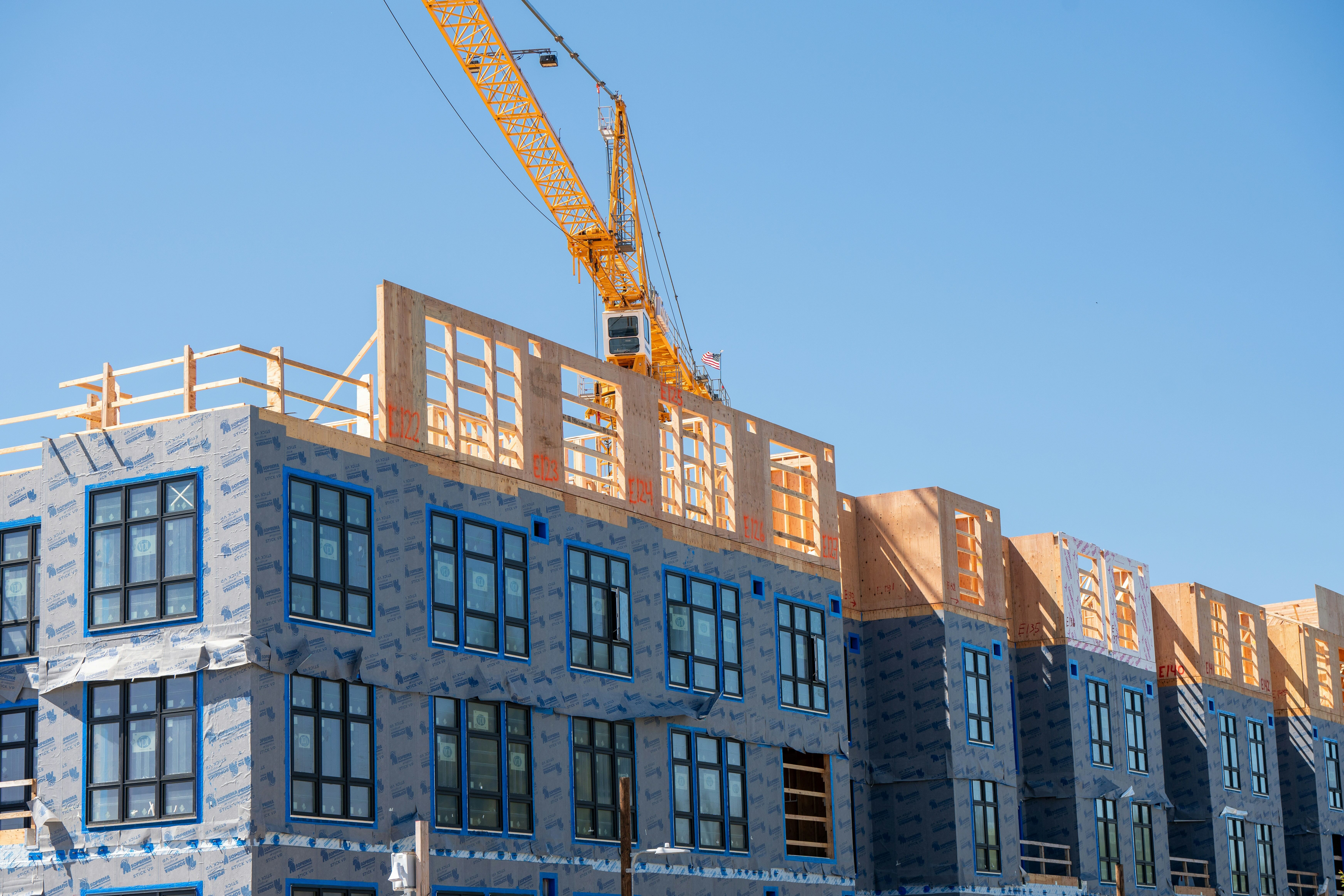 Apartment building under construction with wrapped lower floors and wood framing on top under a blue sky.