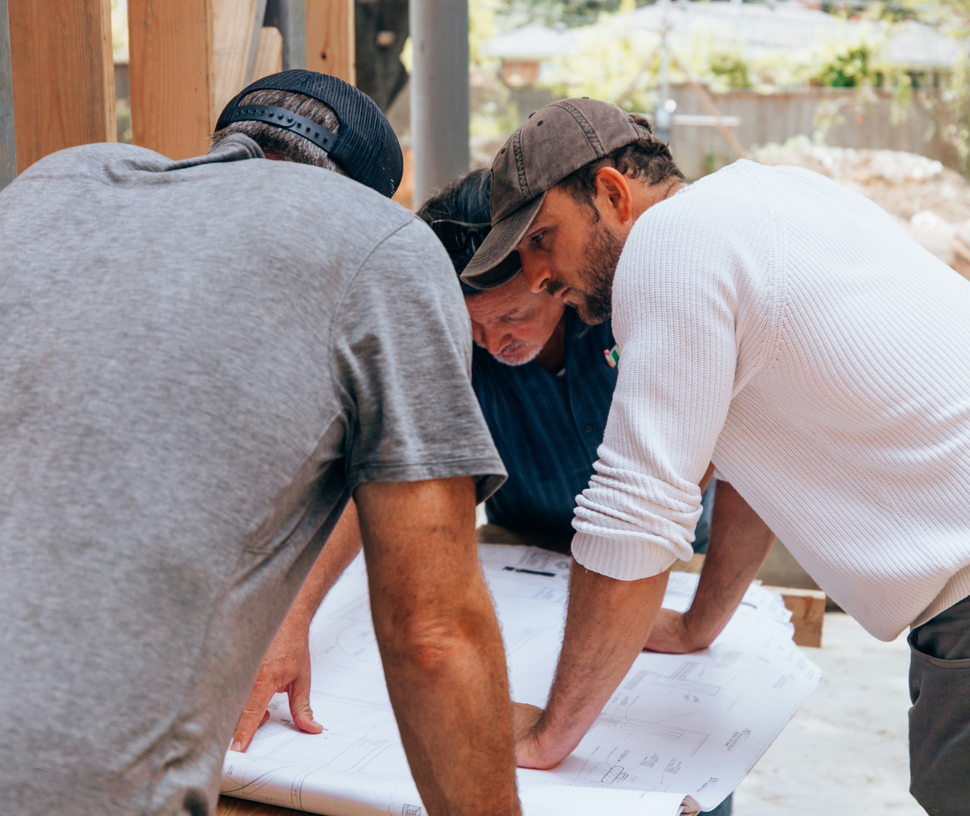 Three construction workers closely examining blueprints on a wooden table at a work site.