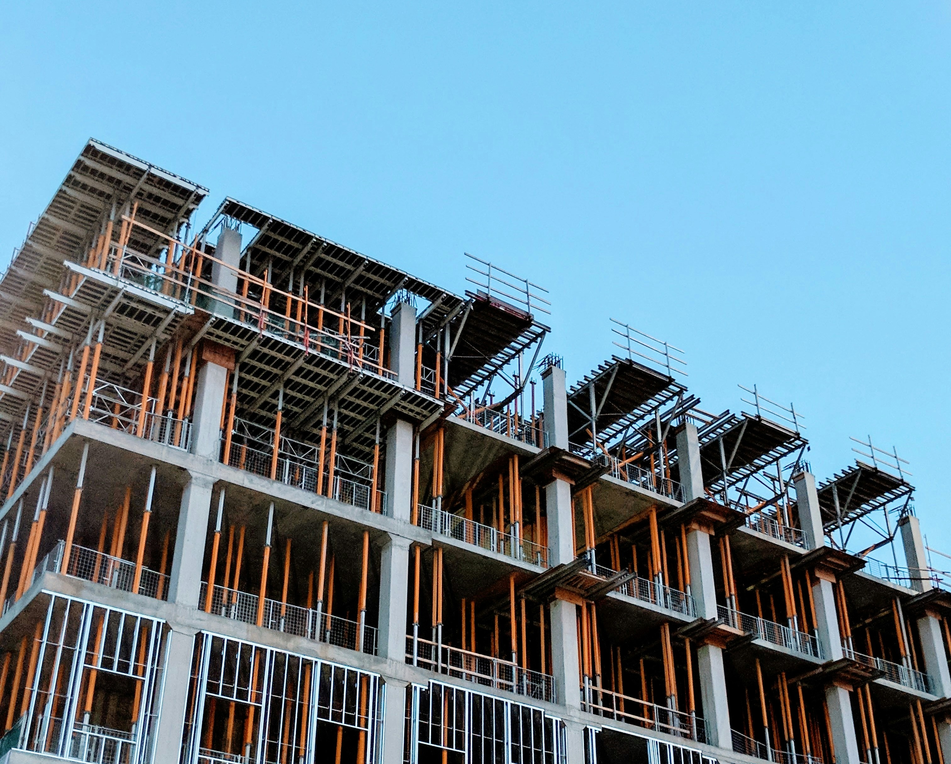 Under-construction multi-story concrete building with scaffolding and support beams against blue sky.
