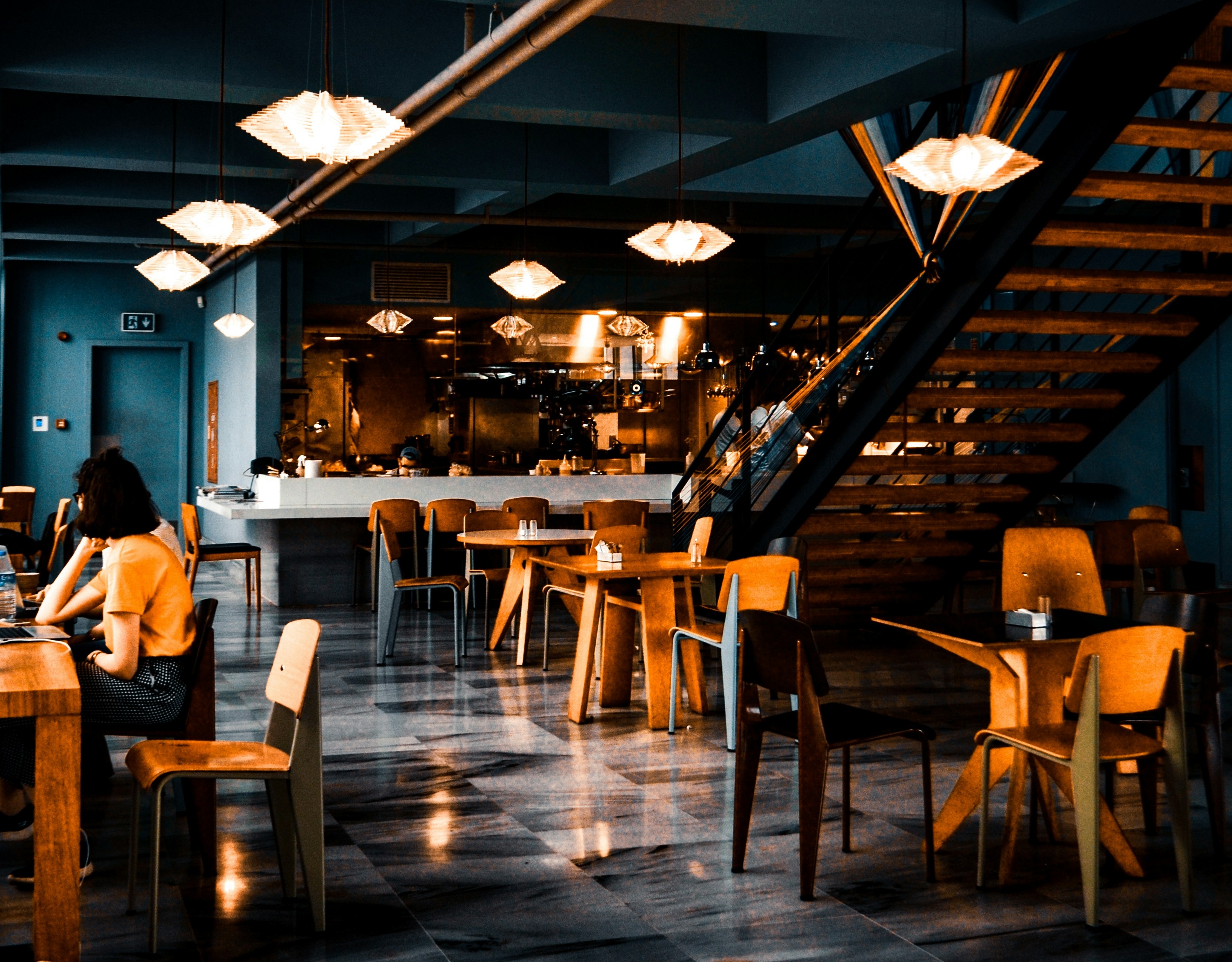 Modern café interior with wooden tables, chairs, hanging lights, and a staircase near the bar.