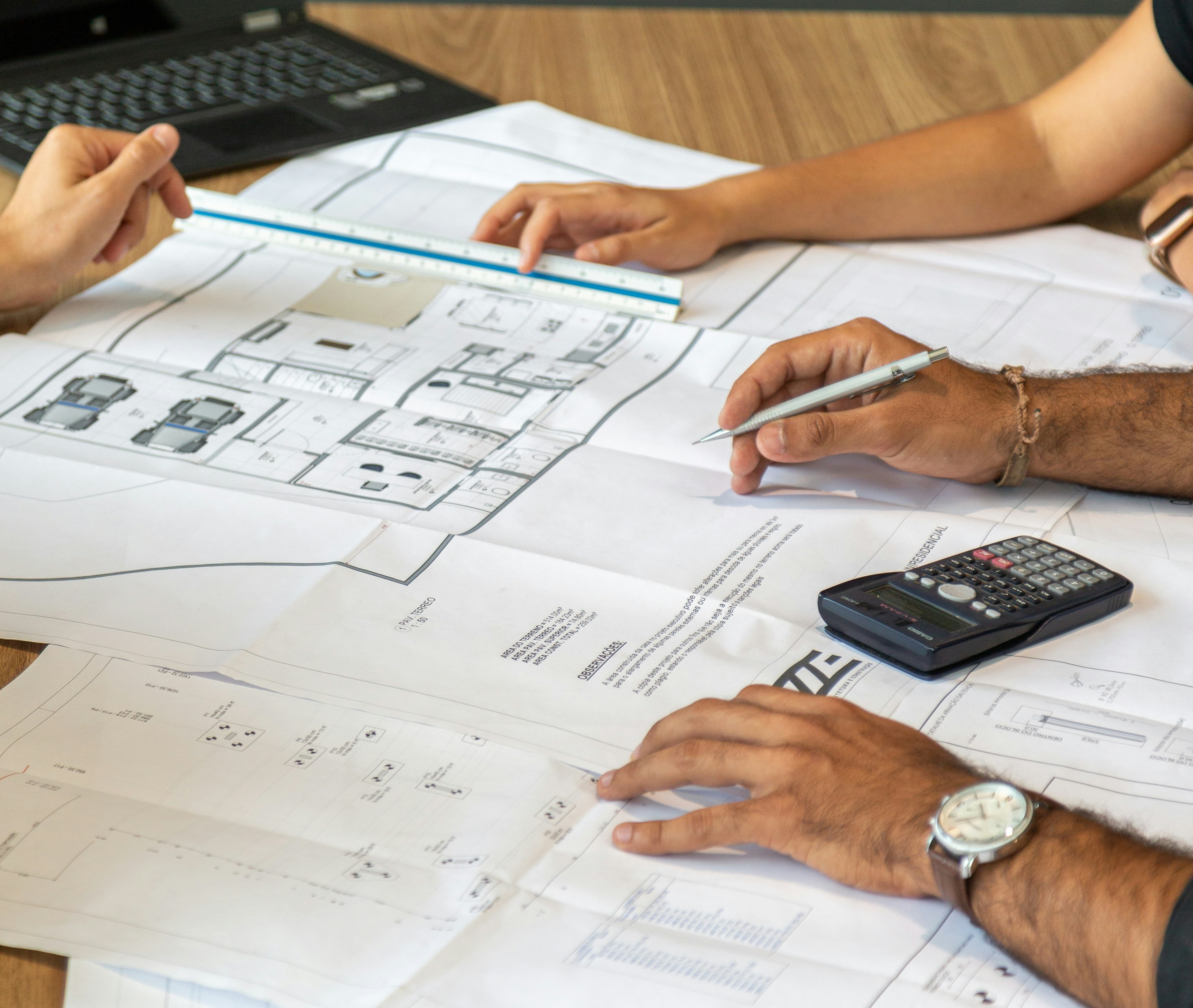 Hands holding pen and ruler over architectural blueprints with calculator on wood table.