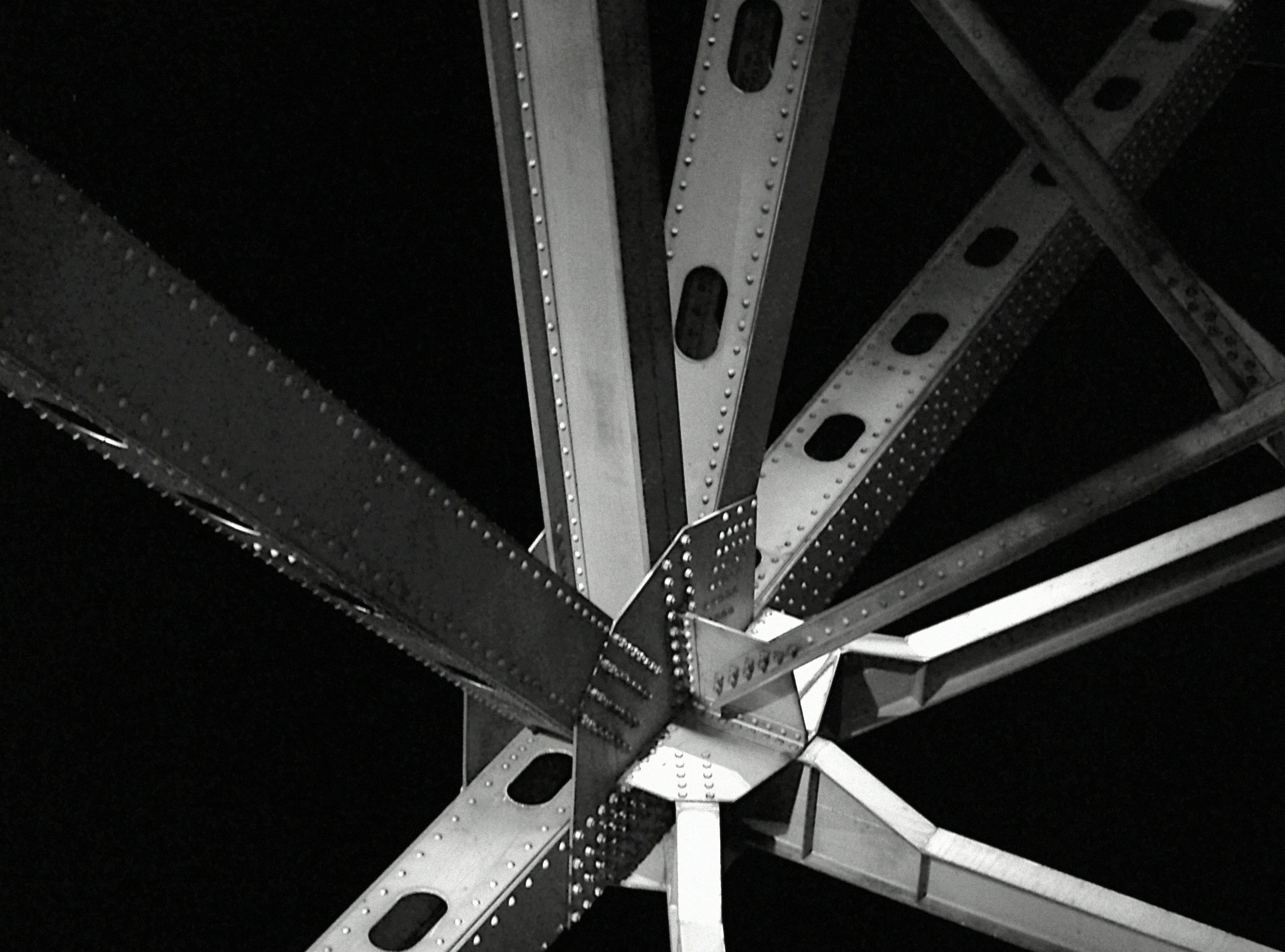 Black and white photo of steel girders joined with rivets forming a triangular structure against black background.