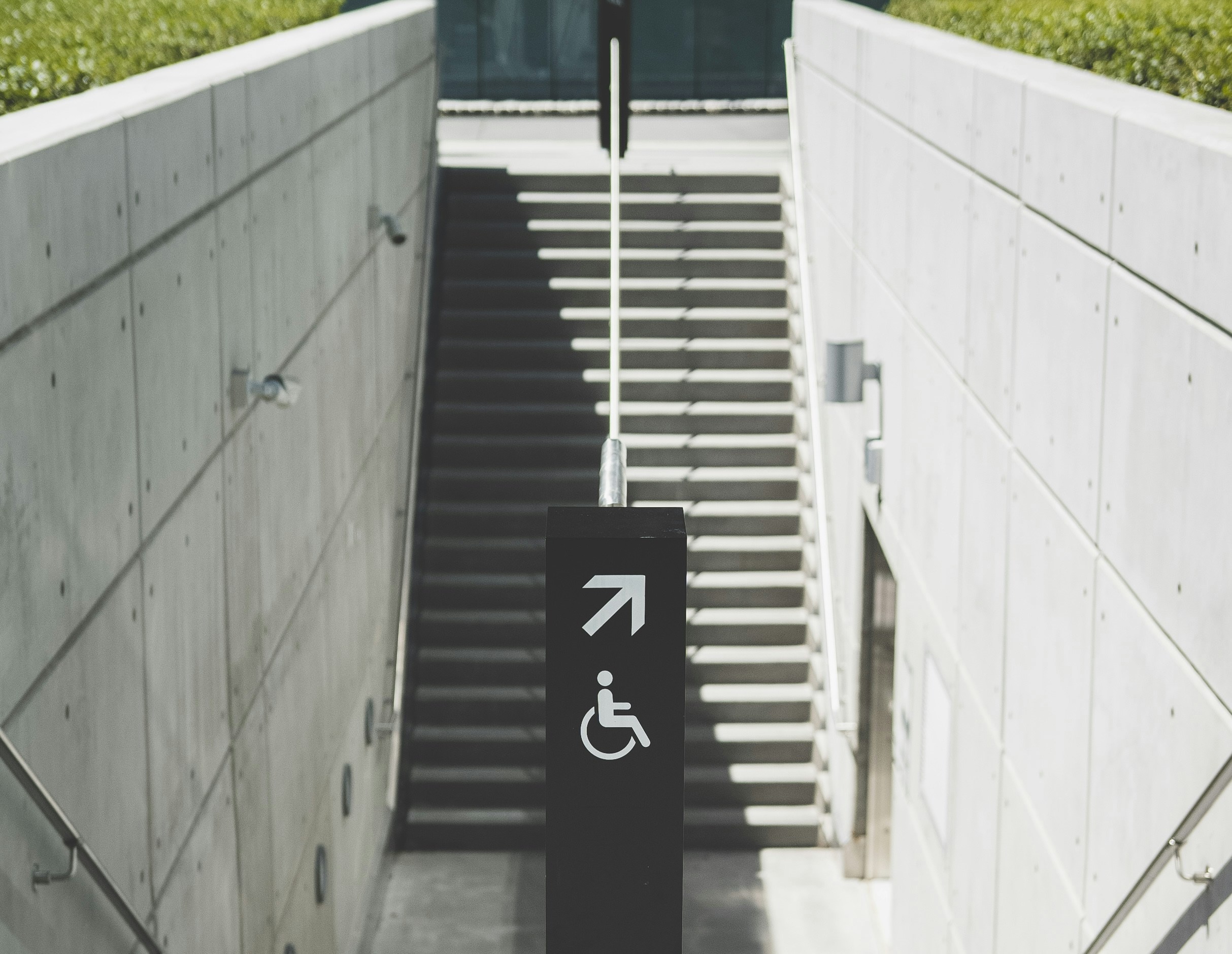 Signpost with a wheelchair symbol and arrow pointing right towards stairs with concrete walls.