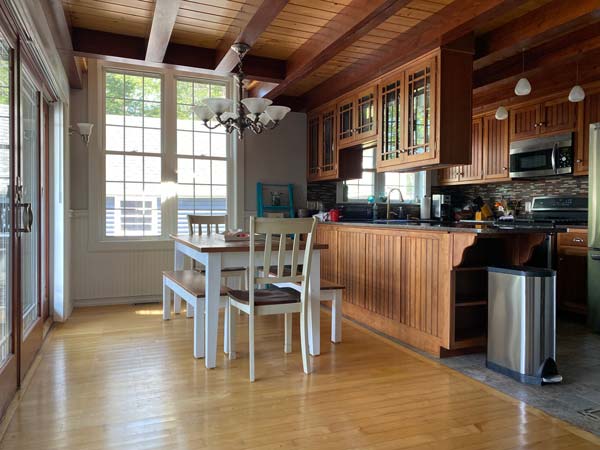 Bright kitchen with wooden cabinets, white dining table, chairs, and large windows letting in natural light.