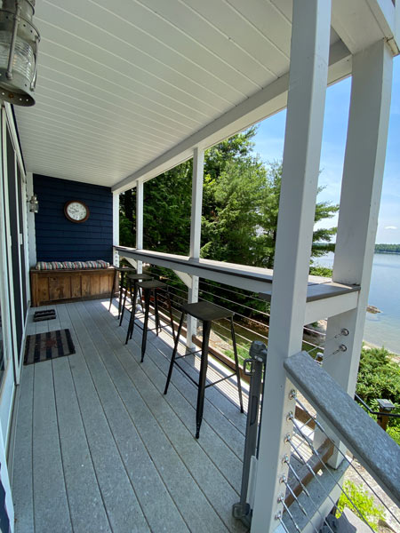 Covered porch with tall chairs along railing overlooking a lake and trees on a sunny day.
