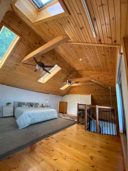 Cozy attic bedroom with wooden ceiling, skylights, ceiling fans, white bed, and wooden floor.