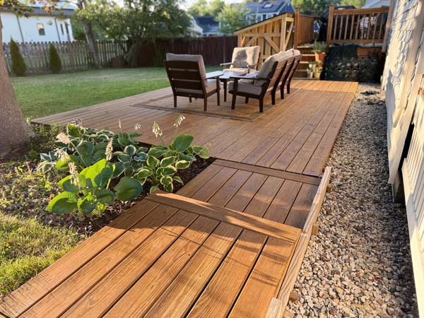 Wooden backyard deck with patio chairs, a table, plants, and a gravel path beside a house.