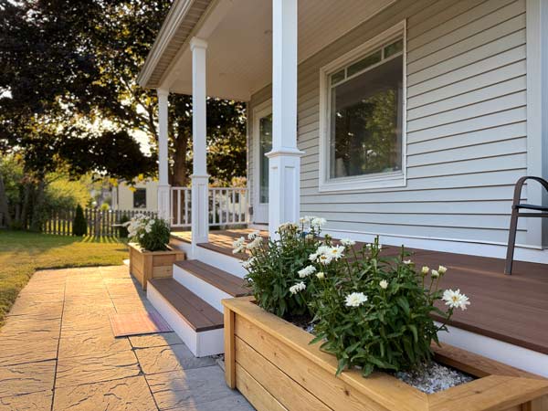 Wooden porch with white columns, steps, and planter boxes filled with white flowers in the late afternoon sun.