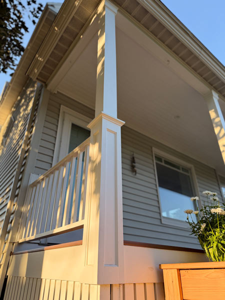 White porch columns supporting a wooden roof with a window reflecting trees.