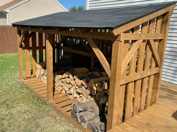 Wooden outdoor shed with stacked firewood inside on a grassy lawn beside houses.