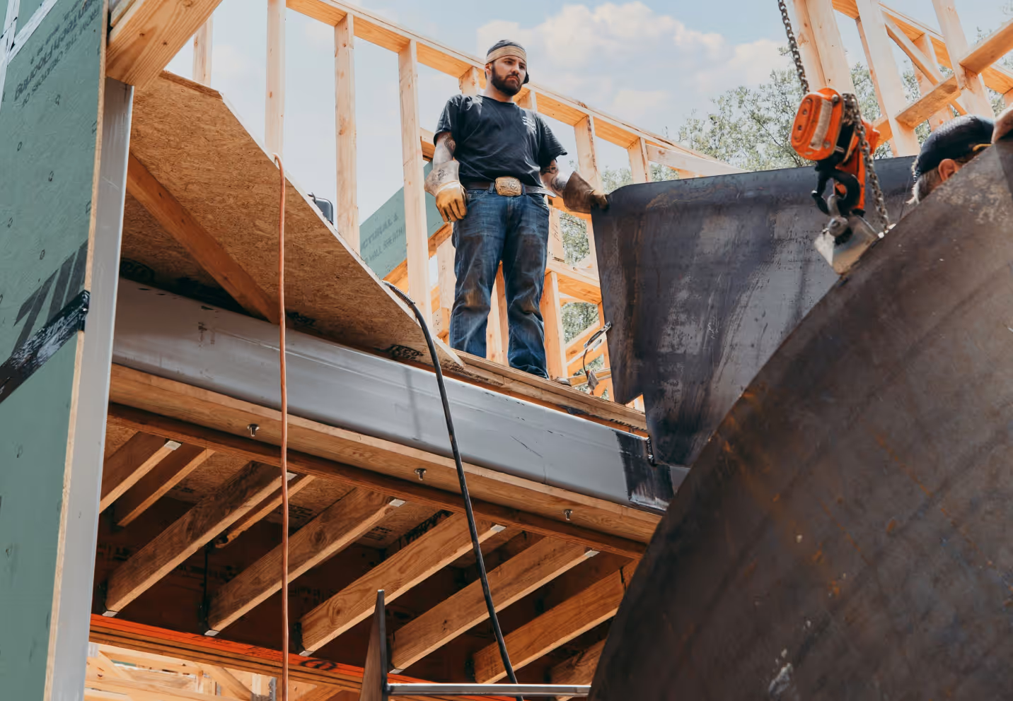 Construction worker in gloves and headband standing on a wooden frame structure guiding a large metal beam being lifted.