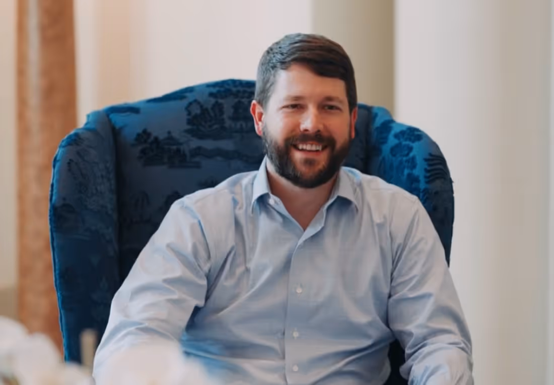 Smiling man with beard sitting in a blue patterned armchair wearing a light blue shirt.