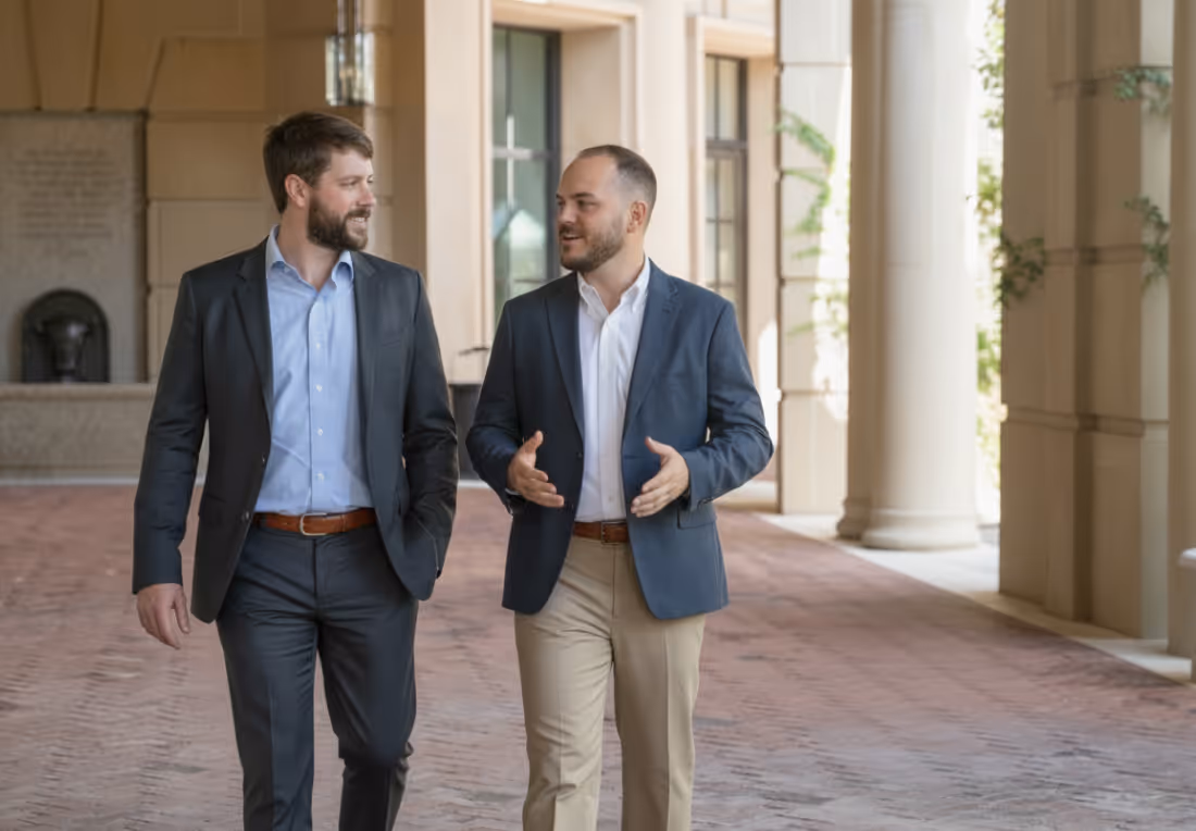 Two men in business attire walking and talking in a corridor with large columns.