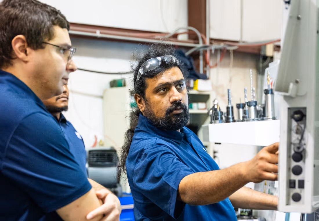 Three factory workers in blue uniforms operating and examining industrial machinery with drill attachments.