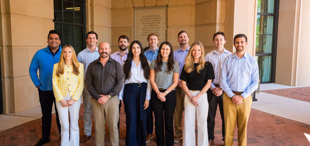 Group of eleven young professionals standing outdoors in front of a building, smiling and dressed in business casual attire.