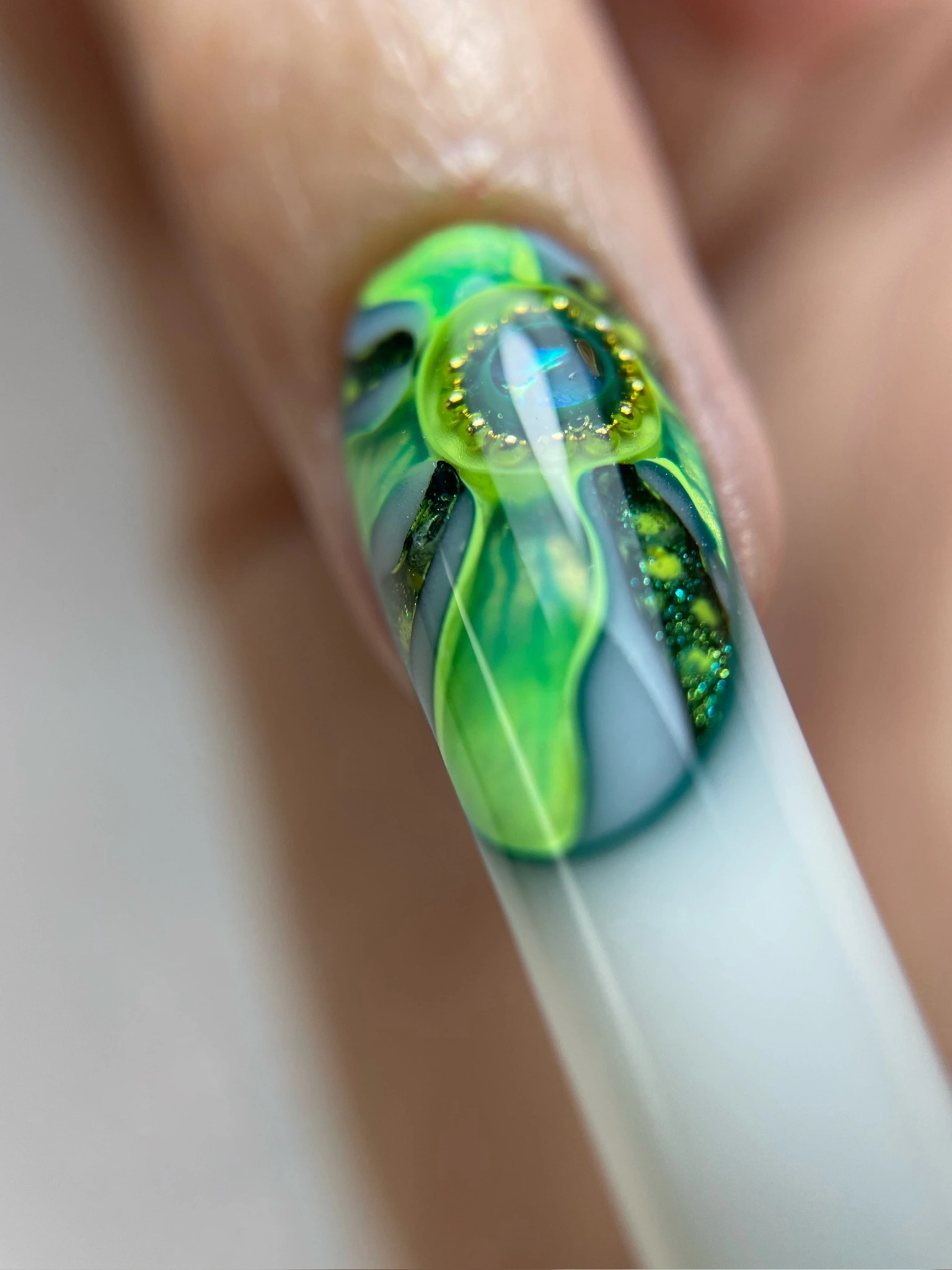 Gros plan sur un ongle orné d'un nail art en gel 3D vert et bleu, représentant une forme ovale en relief rehaussée de perles dorées.