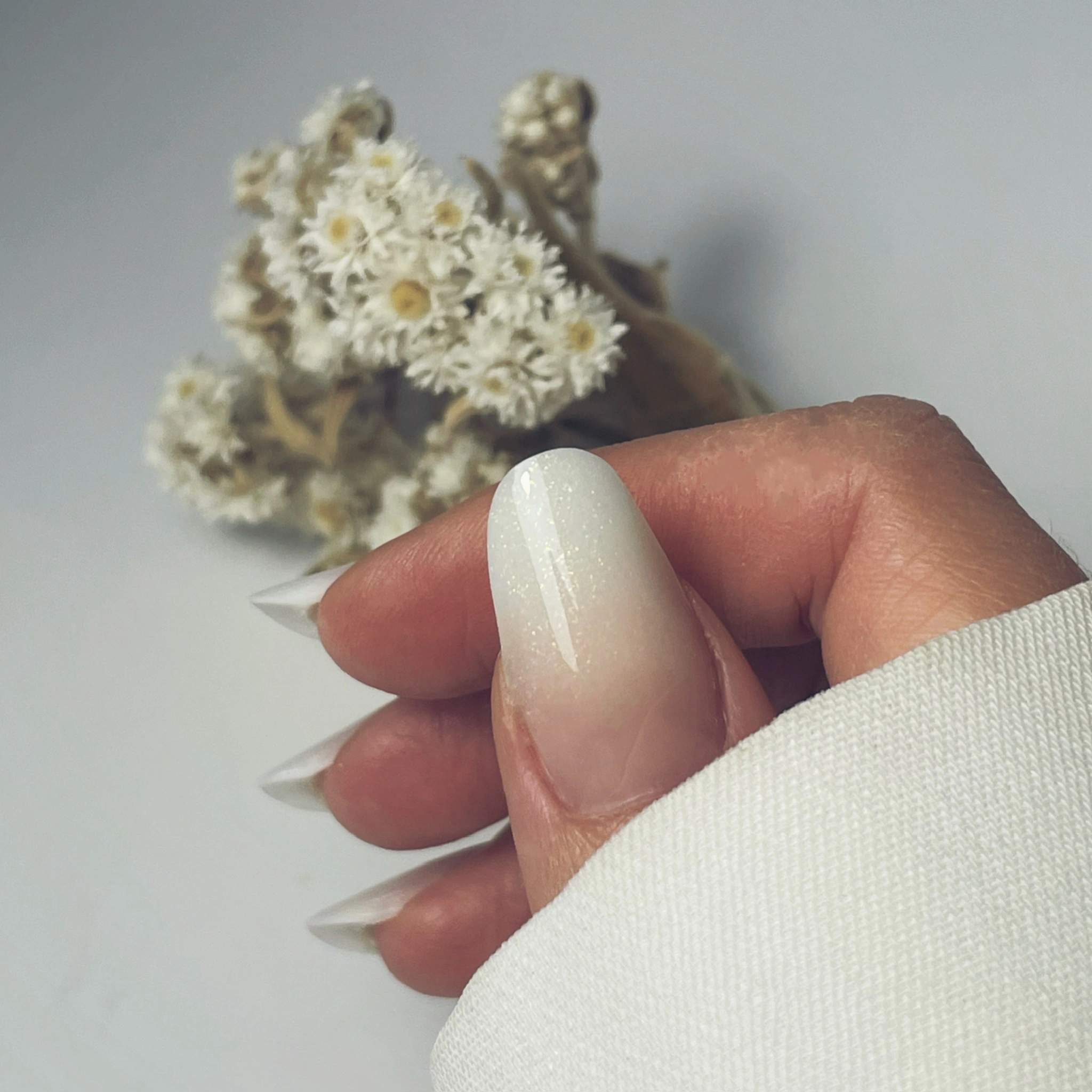 Gros plan sur une main arborant une manucure ombrée blanche pailletée, tenant de petites fleurs séchées blanches.
