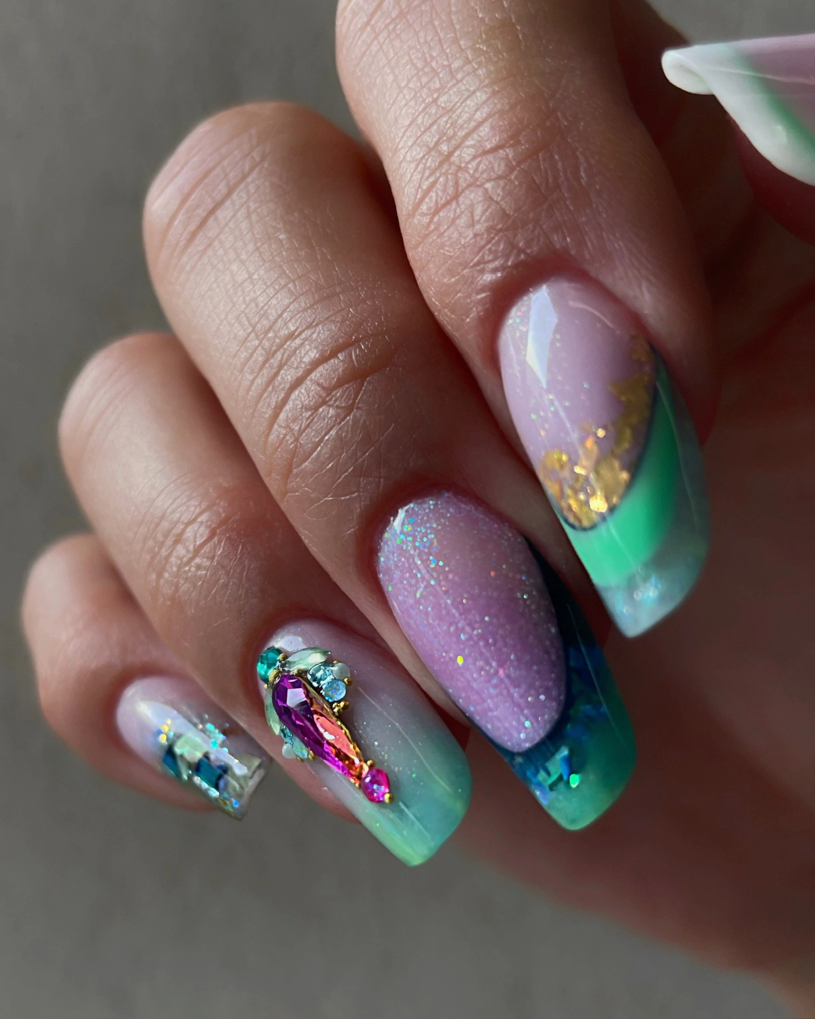 Gros plan sur une main aux ongles longs, ornée d'un nail art coloré comprenant des paillettes, des paillettes dorées et des strass.