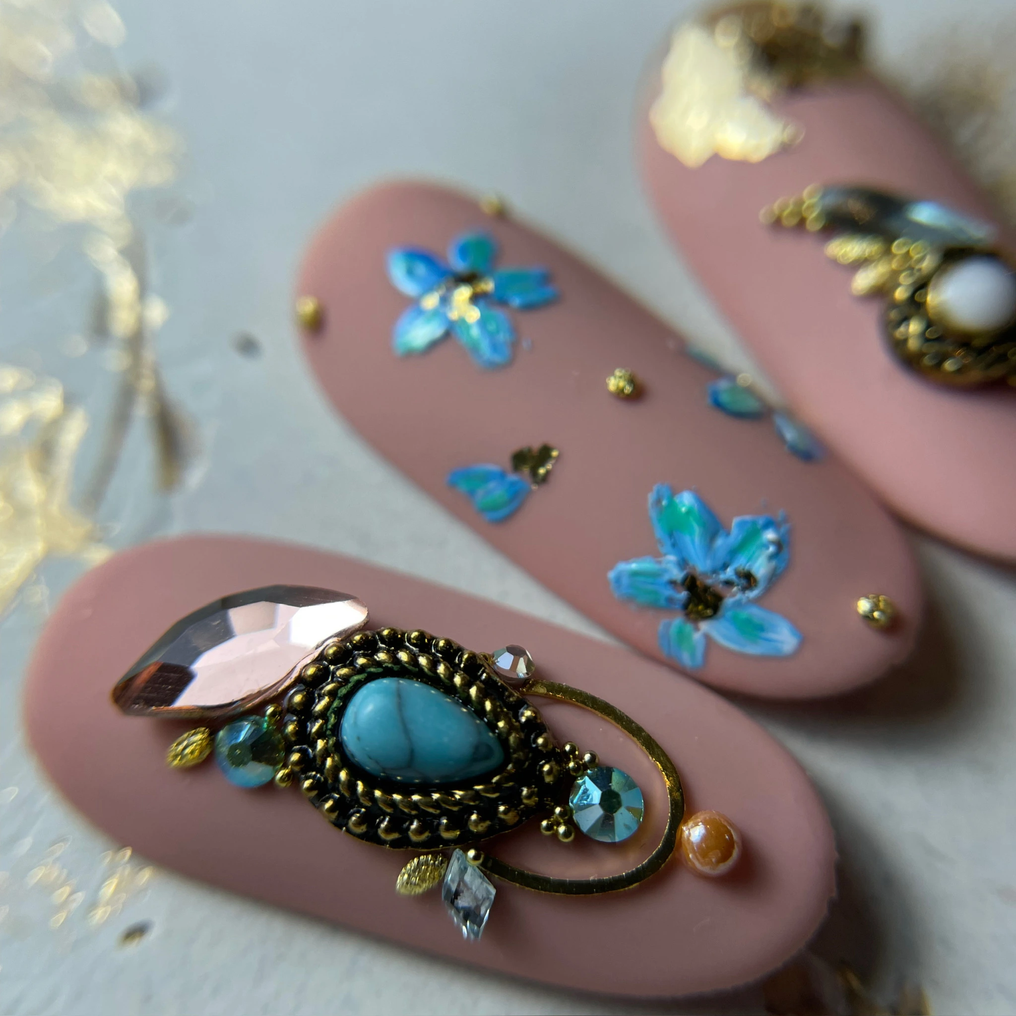 Gros plan sur des ongles artificiels rose mat, ornés de fleurs bleues et de motifs complexes sertis de pierres précieuses dorées et turquoise.