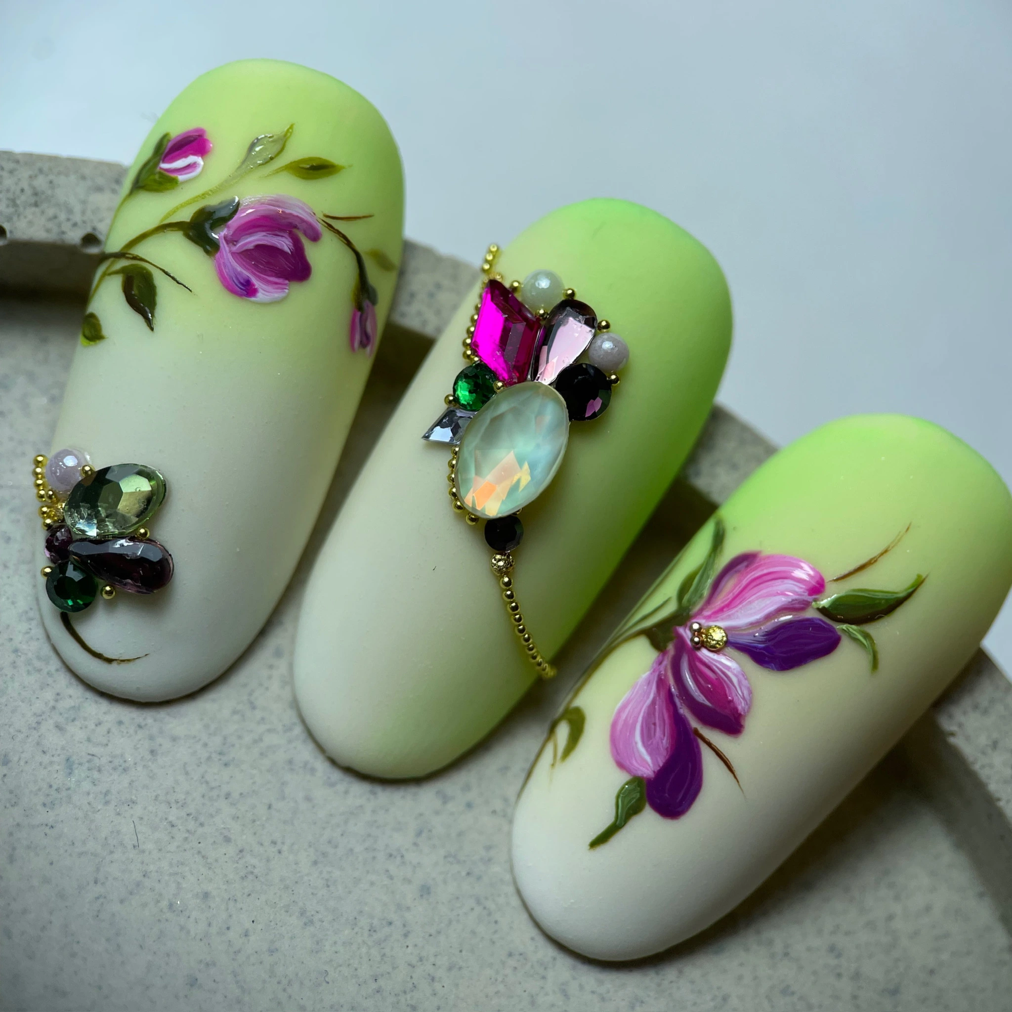 Trois ongles artificiels avec un dégradé vert clair et crème, ornés de motifs floraux roses et violets et de strass colorés.