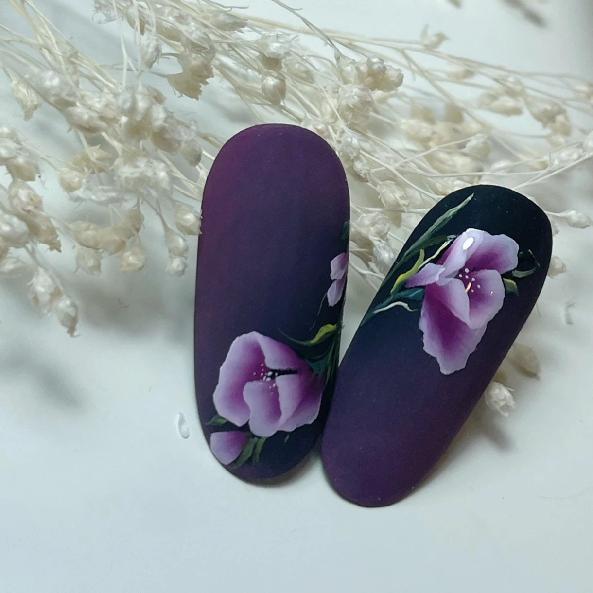 Deux faux ongles dégradés violets, ornés de fleurs violettes et blanches peintes à la main ainsi que de tiges vertes, posés sur un fond blanc avec des fleurs séchées beiges en arrière-plan.