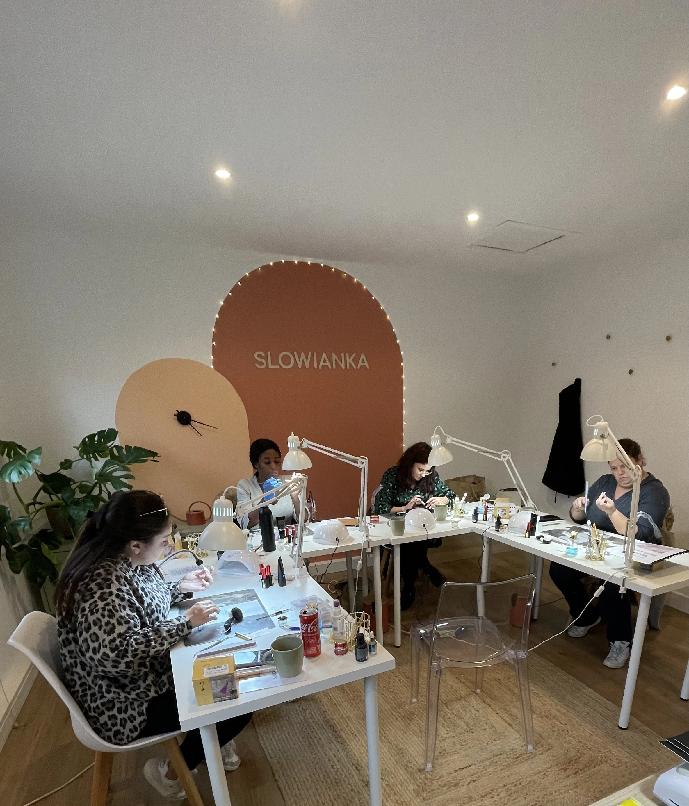 Quatre femmes assises à des tables blanches dans un salon de manucure appelé SLOWIANKA, concentrées sur leur travail de manucure, avec des lampes et des produits de soin des ongles posés sur les tables.