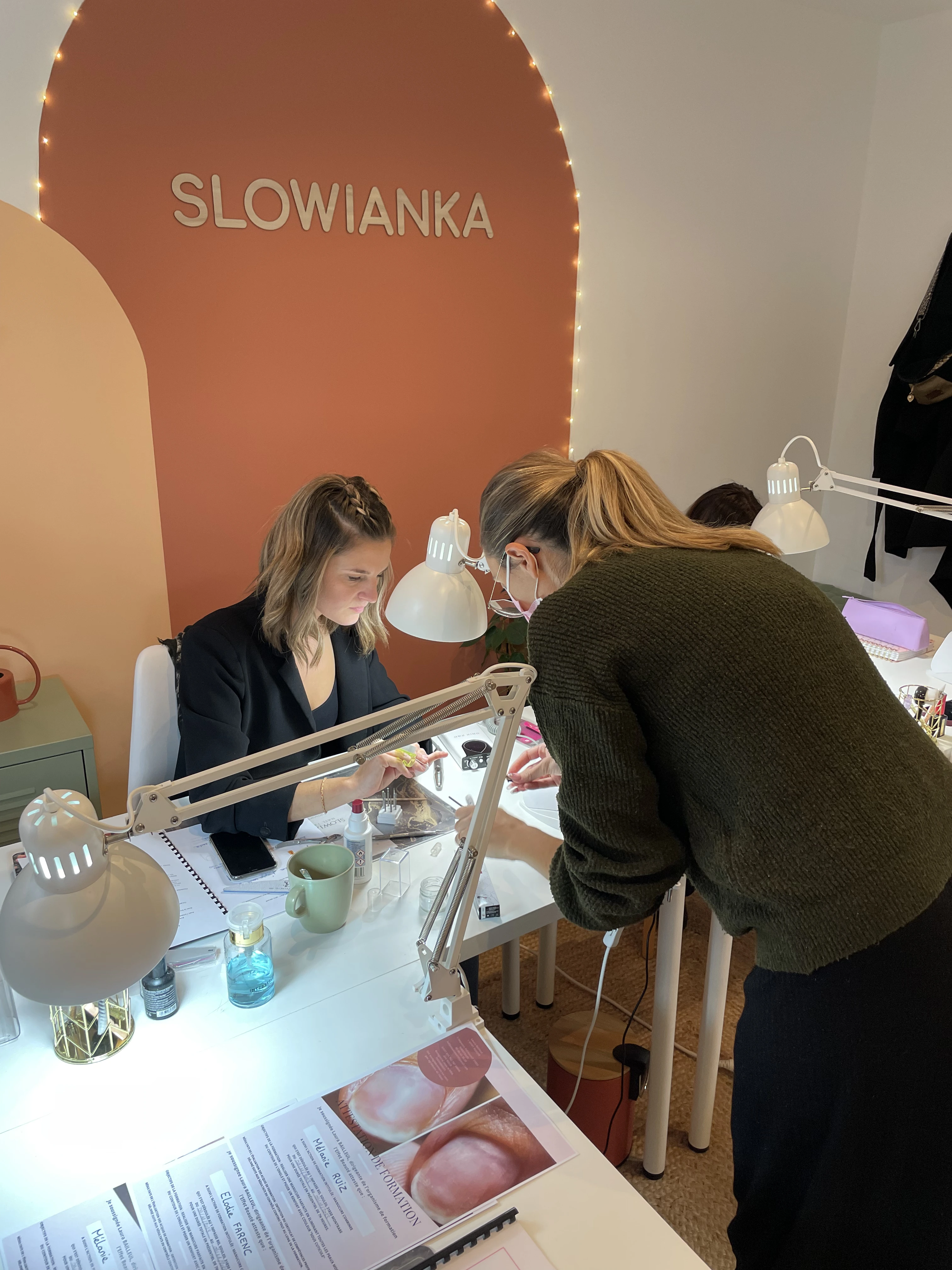 Deux femmes assises à une table de salon de manucure équipée de lampes à UV, l'une s'occupant des ongles tandis que l'autre se penche pour l'aider, avec une enseigne « SLOWIANKA » accrochée au mur.