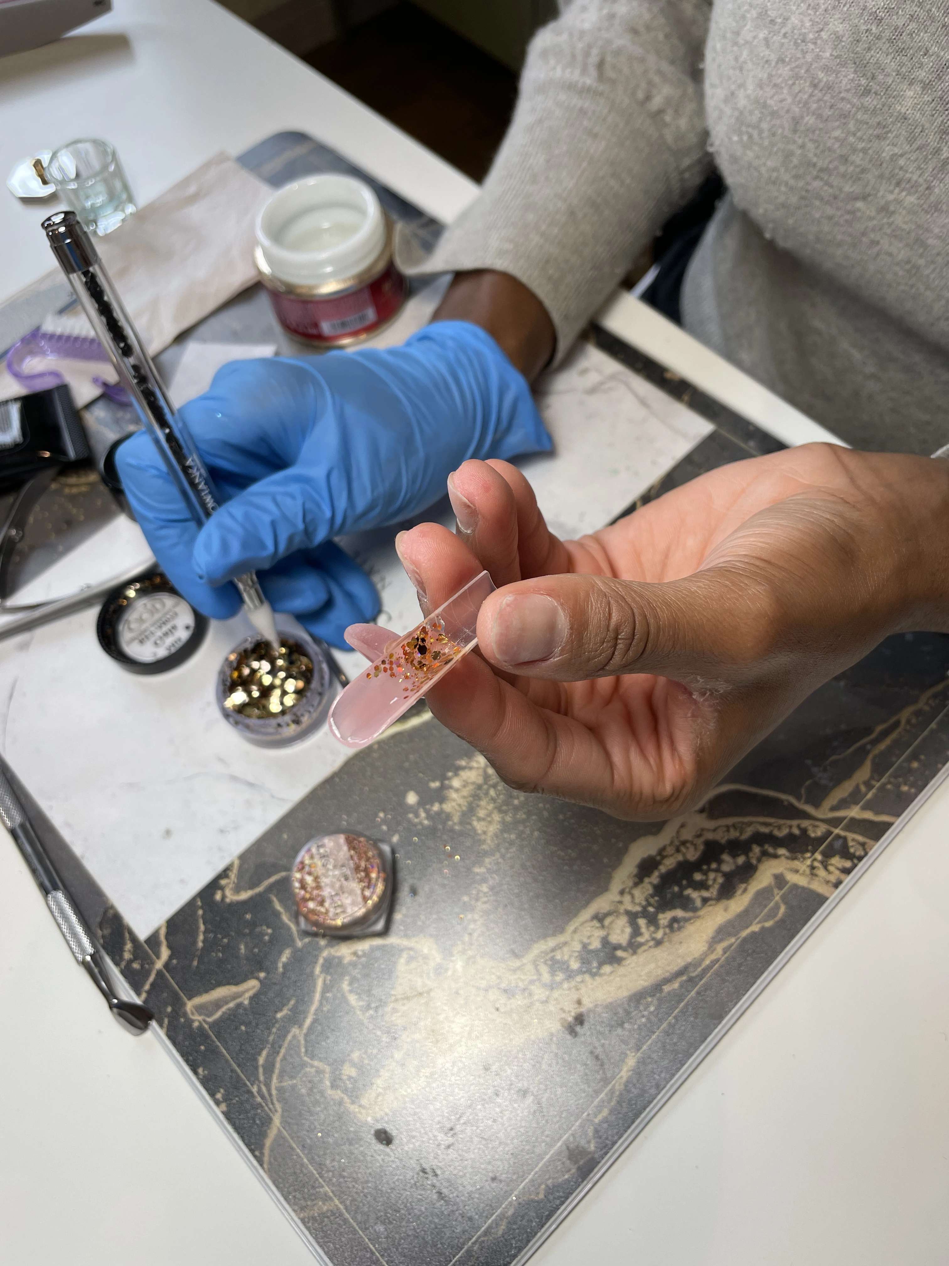 Des mains en train de réaliser une manucure décorative en acrylique avec des paillettes à l'aide d'un stylo à pois, dans un salon de manucure.