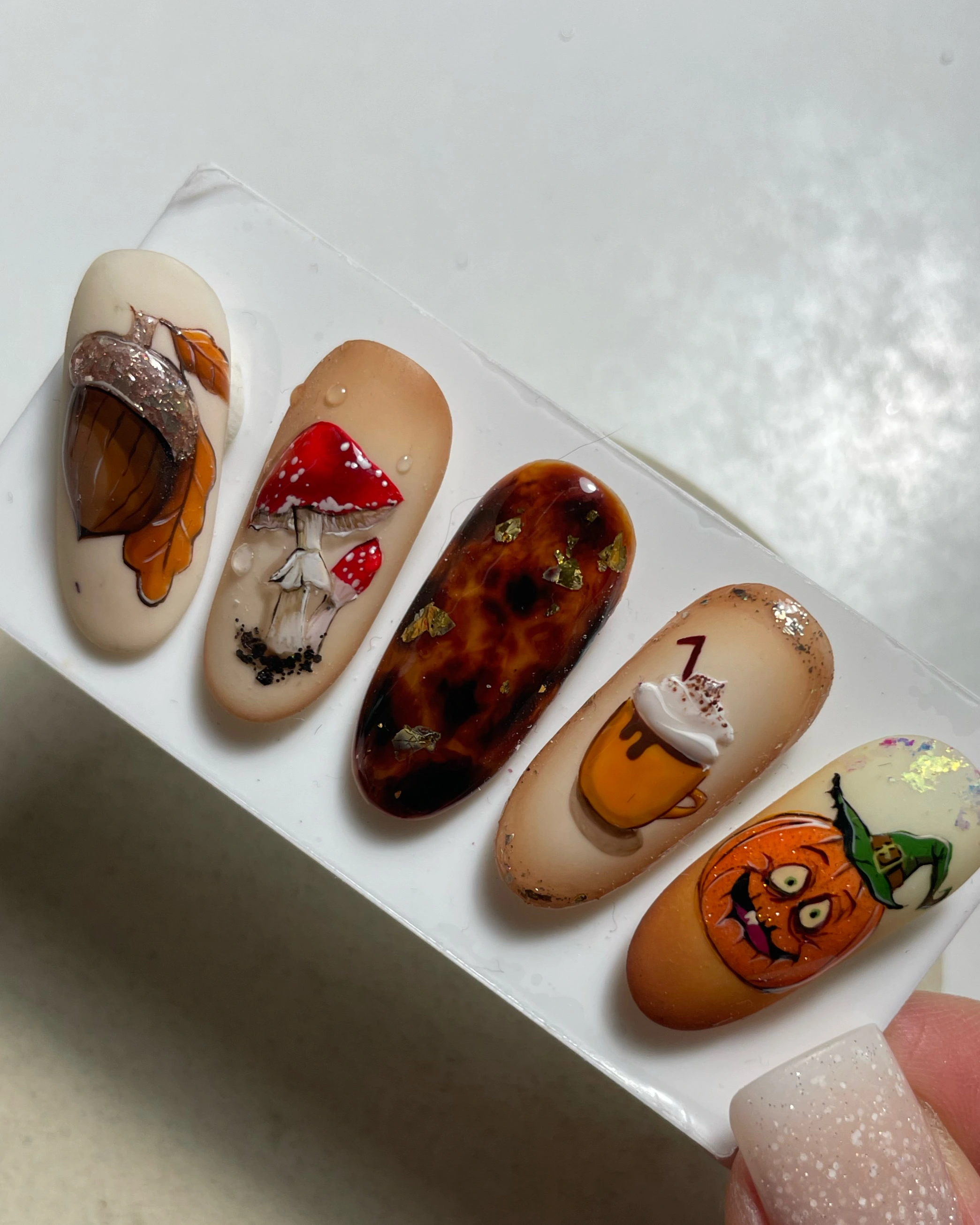 Lot de cinq faux ongles aux motifs automnaux, comprenant notamment un gland orné de feuilles orange, des champignons rouges, un motif écaille de tortue, un latte à la citrouille épicée et une citrouille coiffée d'un chapeau de sorcière vert.