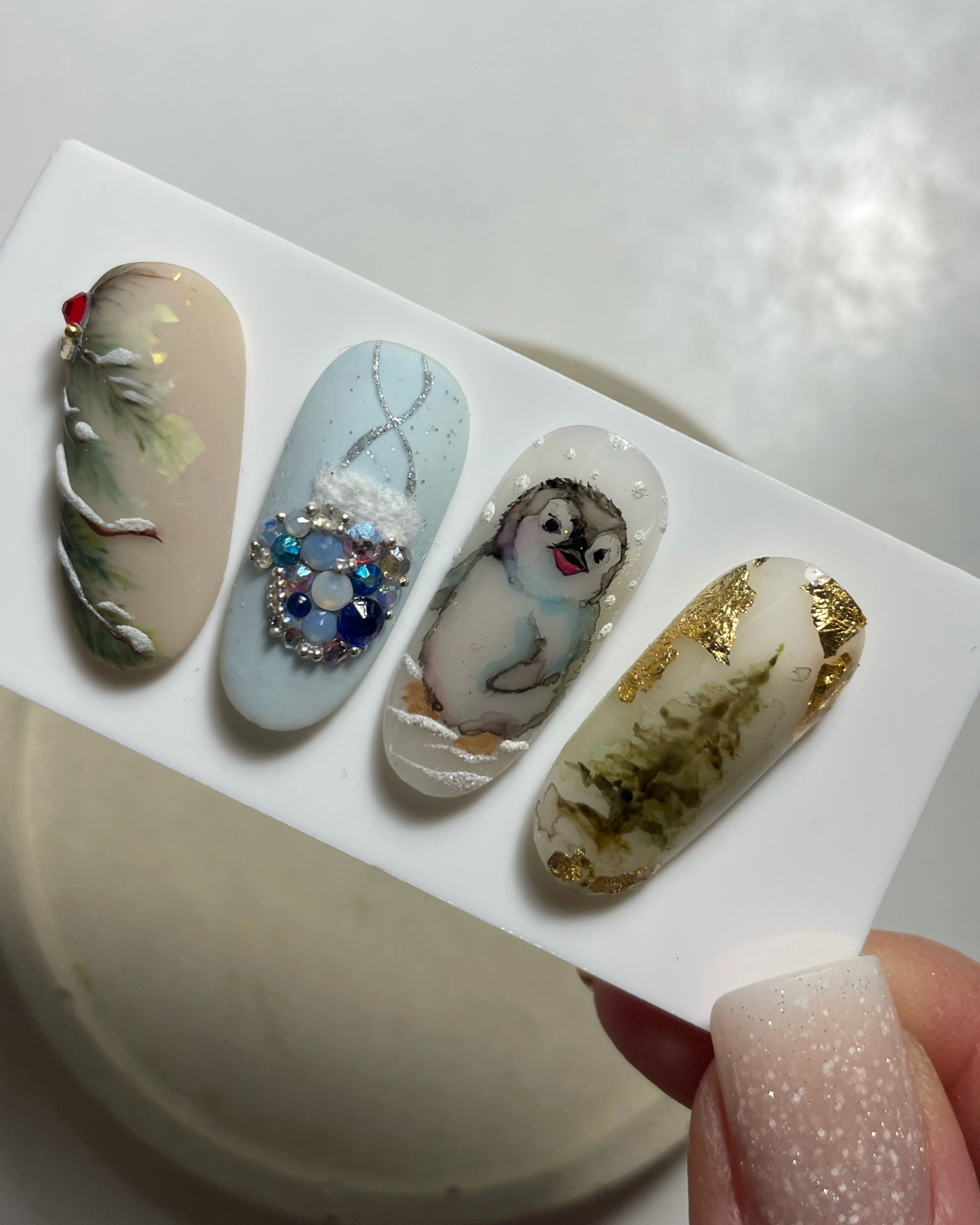 Kit de nail art sur le thème de l'hiver comprenant une branche enneigée ornée d'un bijou rouge, un ongle bleu parsemé de strass, un adorable pingouin dans la neige et un sapin rehaussé de touches dorées.