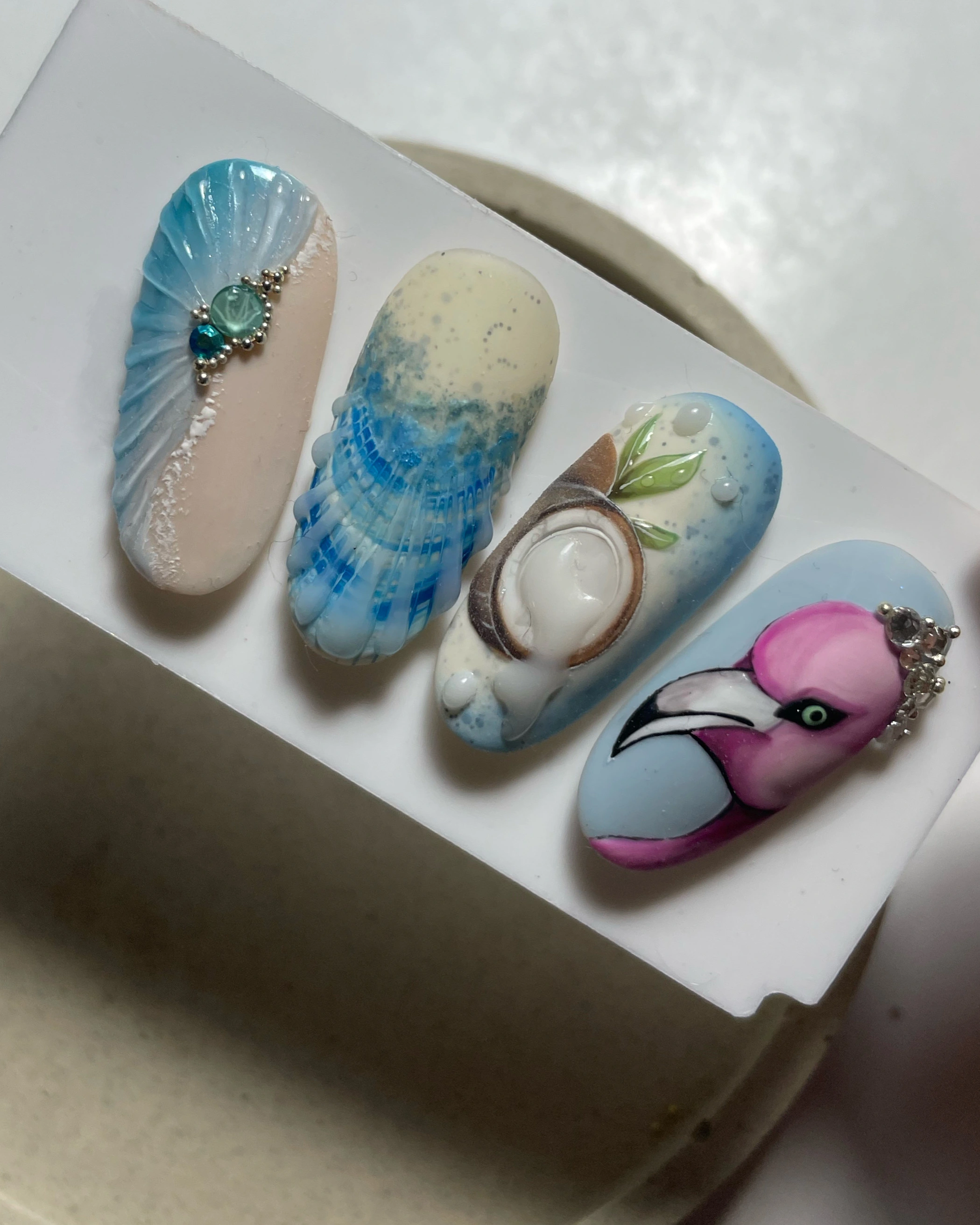 Quatre motifs détaillés pour ongles en acrylique : un coquillage bleu orné de pierres précieuses, une méduse, une noix de coco avec des feuilles et des gouttelettes d'eau, ainsi qu'un flamant rose parsemé de strass.
