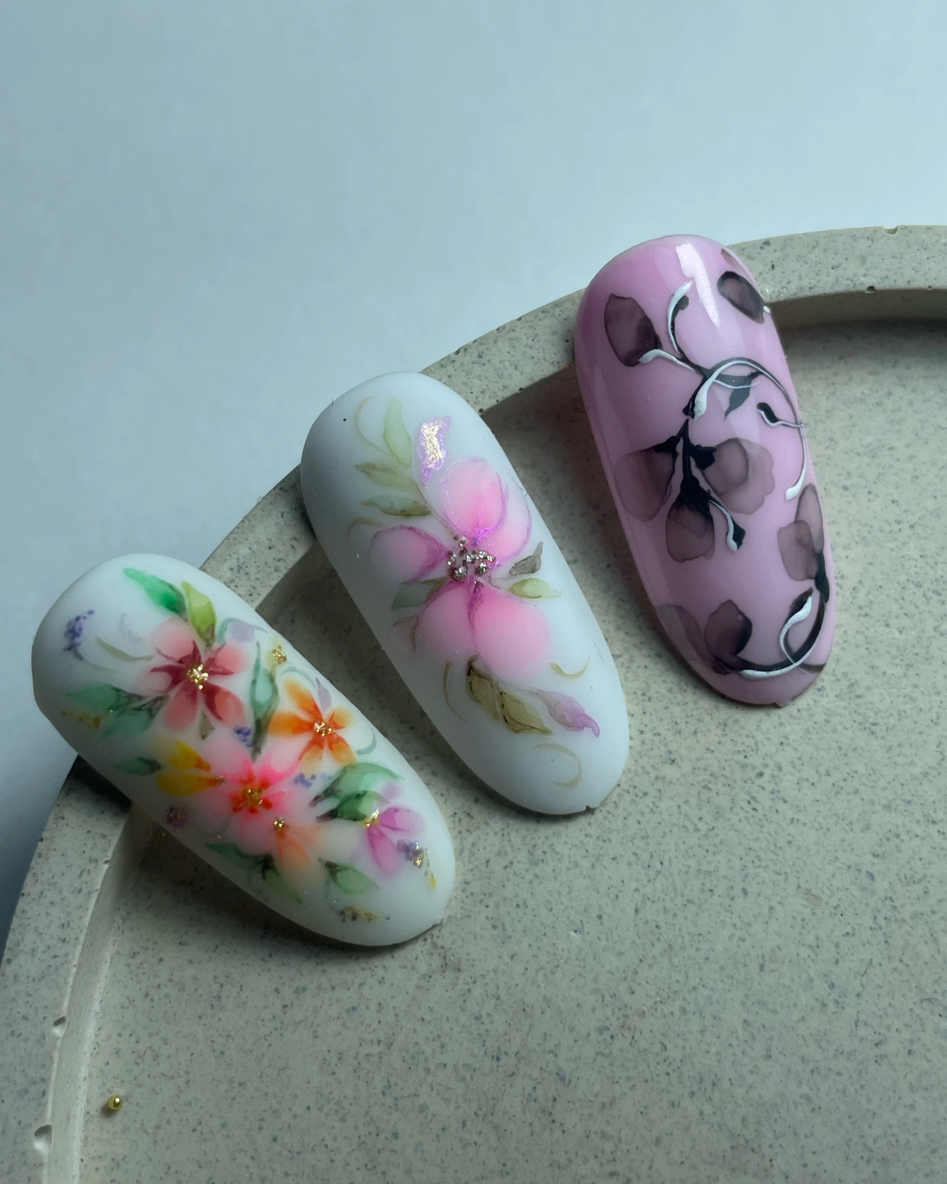 Trois ongles artificiels ornés d'un délicat motif floral en rose, orange et noir sur fond blanc et lavande.
