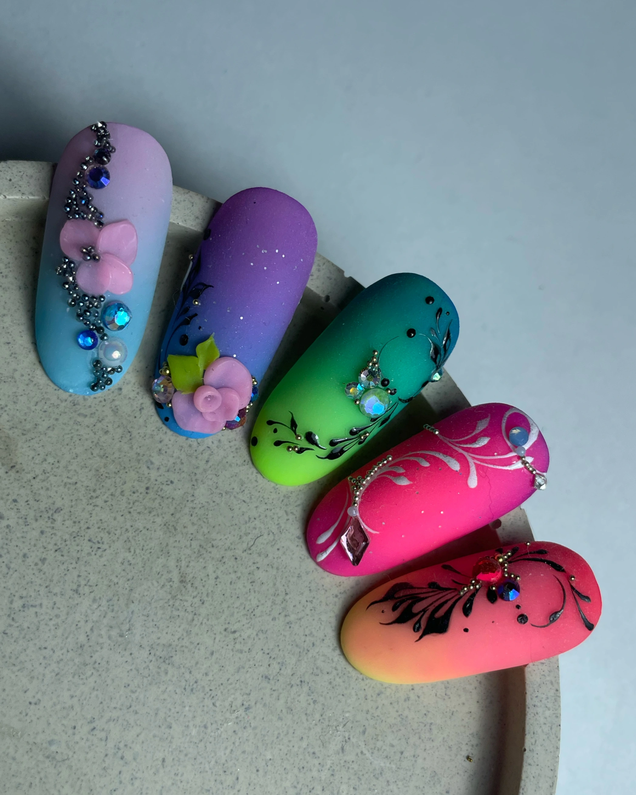 Cinq ongles artificiels présentant un dégradé de couleurs et des motifs floraux en 3D très travaillés, agrémentés de strass, sur une surface ronde de couleur grise.