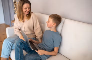 Mother and son sitting on a white couch, smiling and talking while using a laptop together.