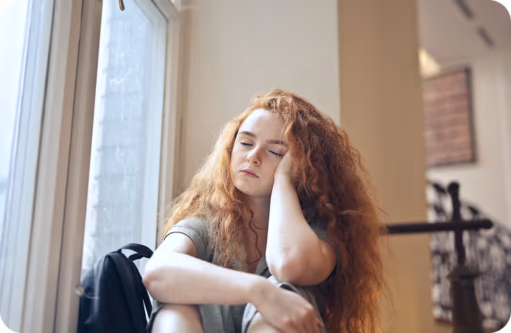 Young woman with long red hair sitting by a window with eyes closed, resting her head on her hand.