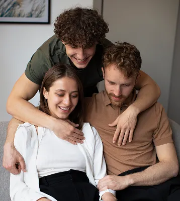 Three friends sitting closely, smiling and hugging each other in a cozy indoor setting.