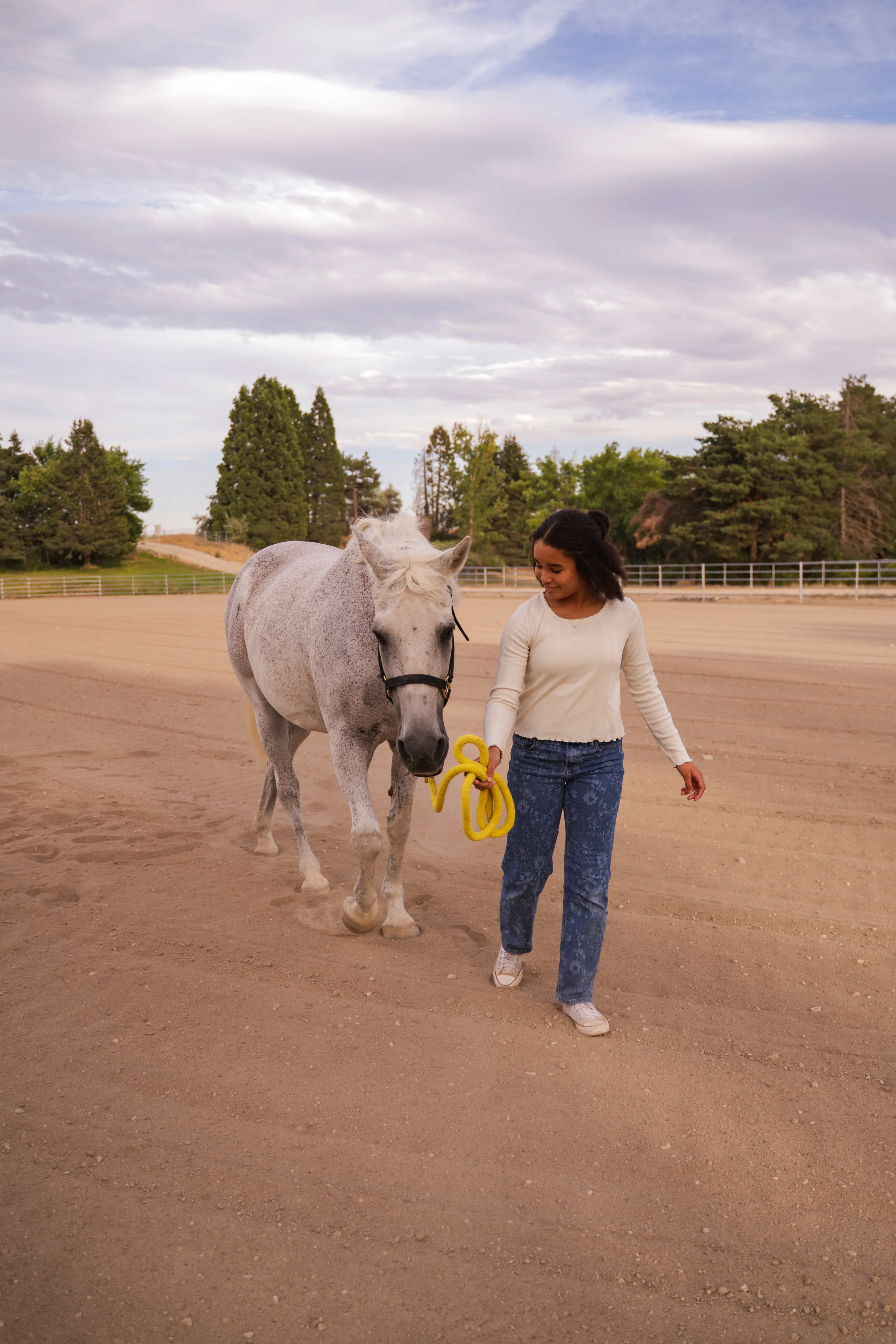 Young woman walking on dirt with a gray horse led by a yellow rope in a fenced area.