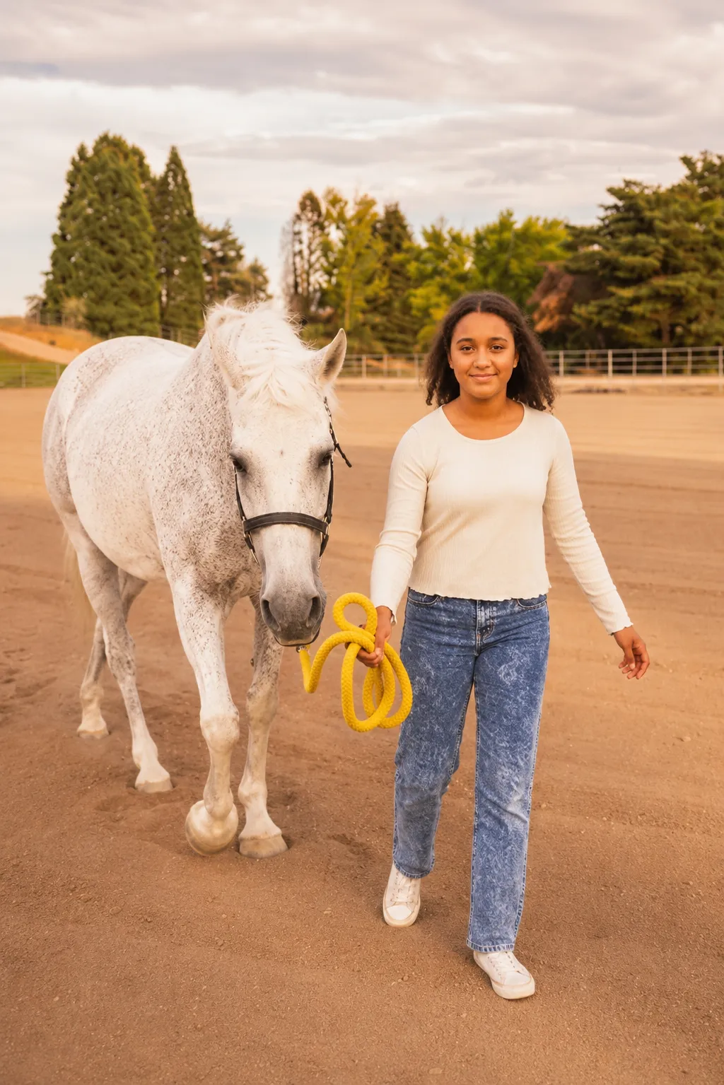 Young woman walking a white horse with a yellow lead rope on a dirt path outdoors.