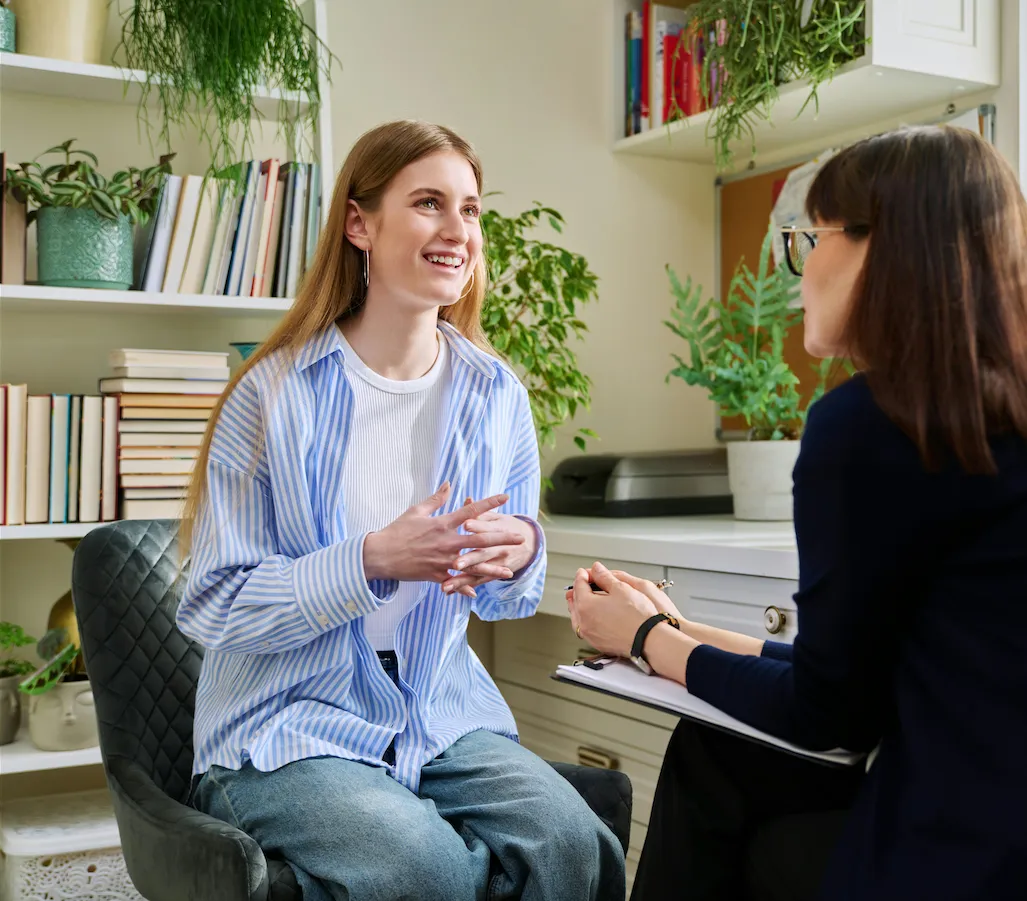 Young woman smiling and talking with a counselor in a cozy office with plants and books.