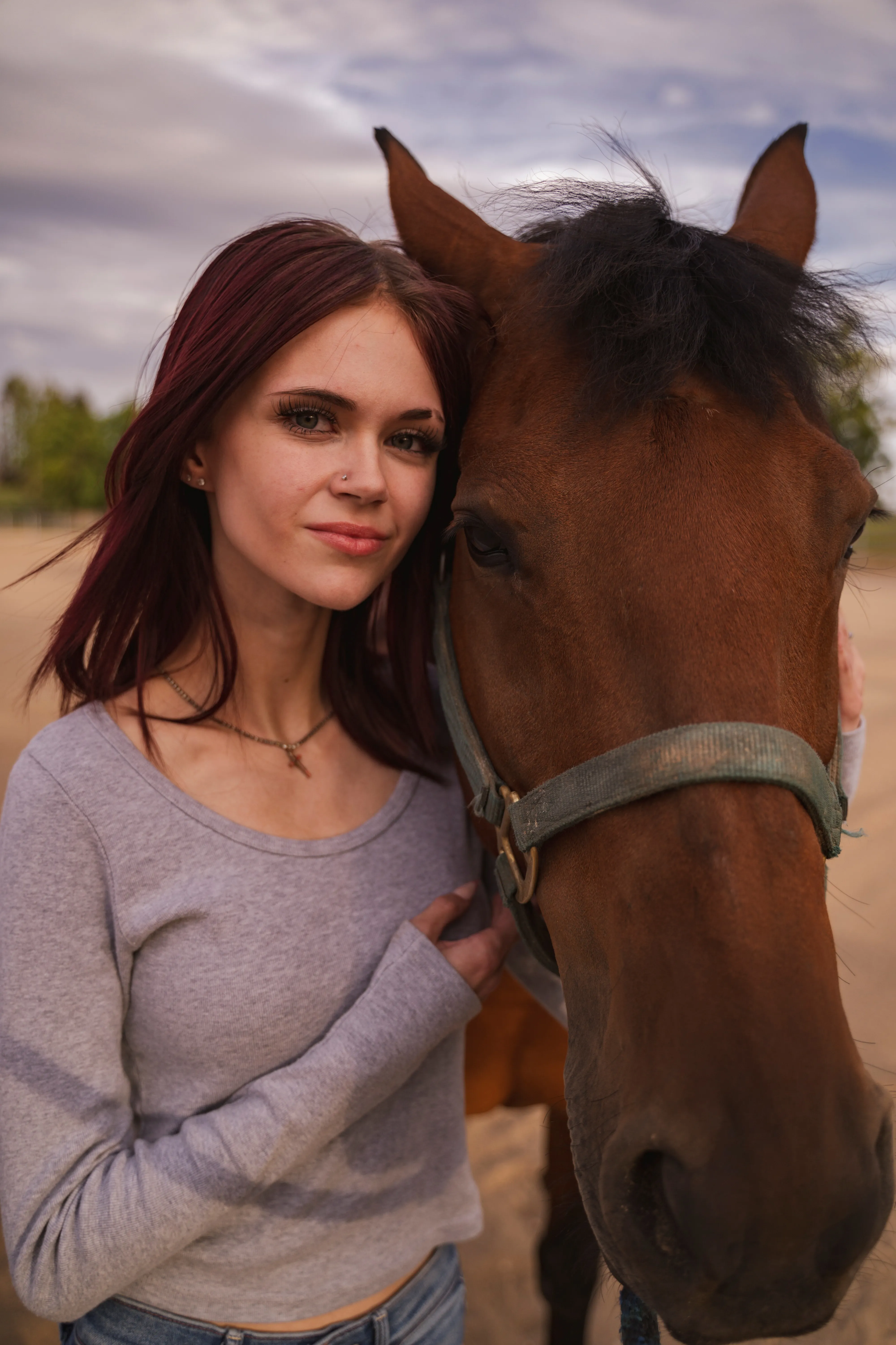 Young woman with red hair standing close to a brown horse, both facing the camera outdoors.