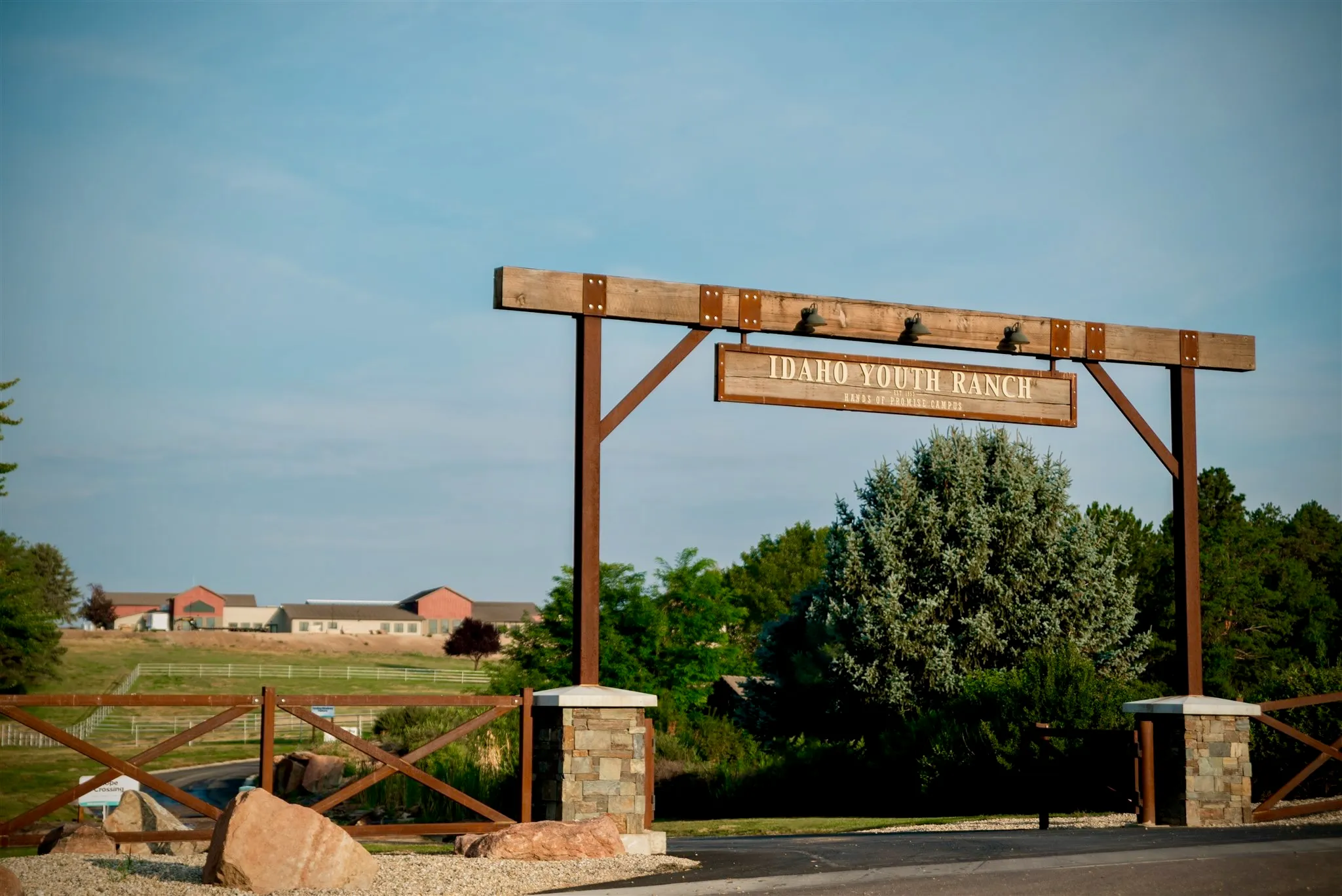 Wooden entrance sign reading Idaho Youth Ranch at a ranch with buildings and trees in the background.