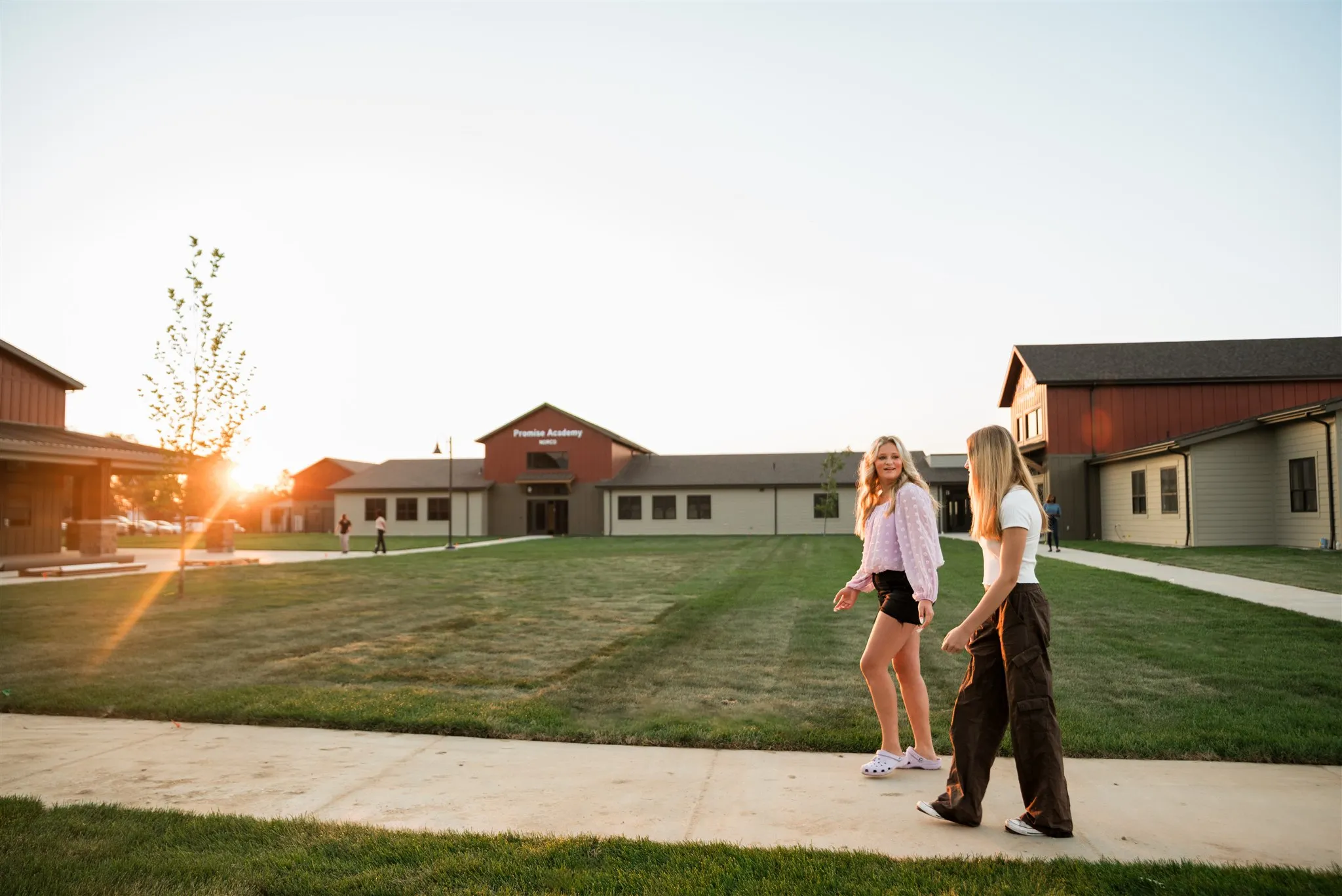 Two young women walk and talk on a concrete path near grassy lawns and school buildings at sunset.